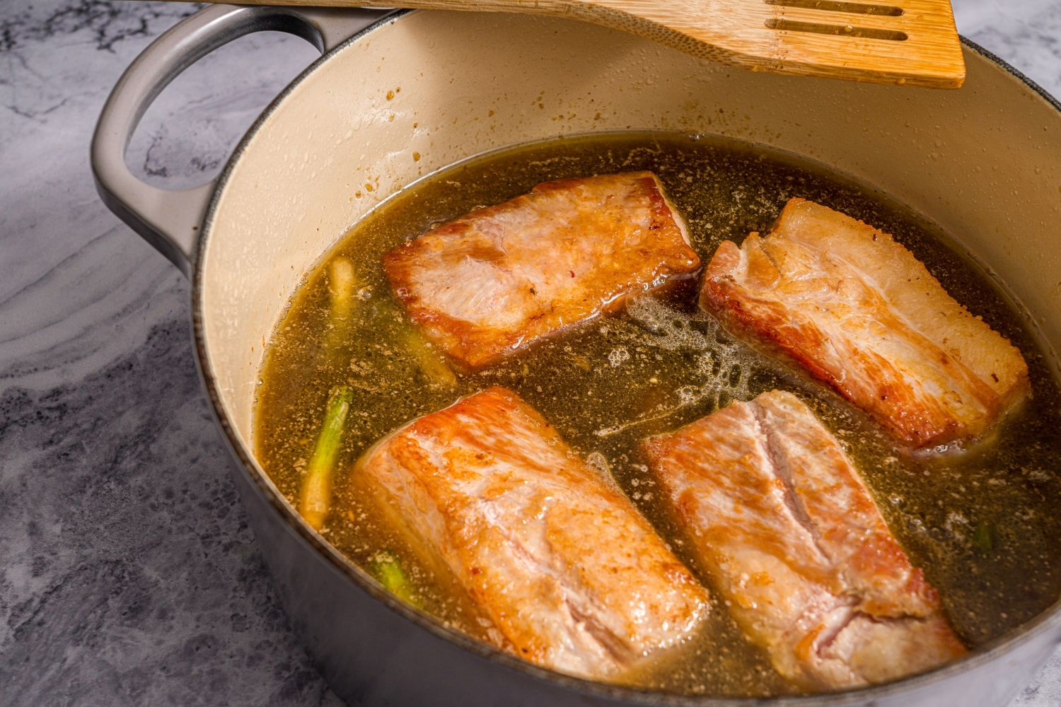 A dutch oven with pork belly braising in broth. The pot is on a marble counter.
