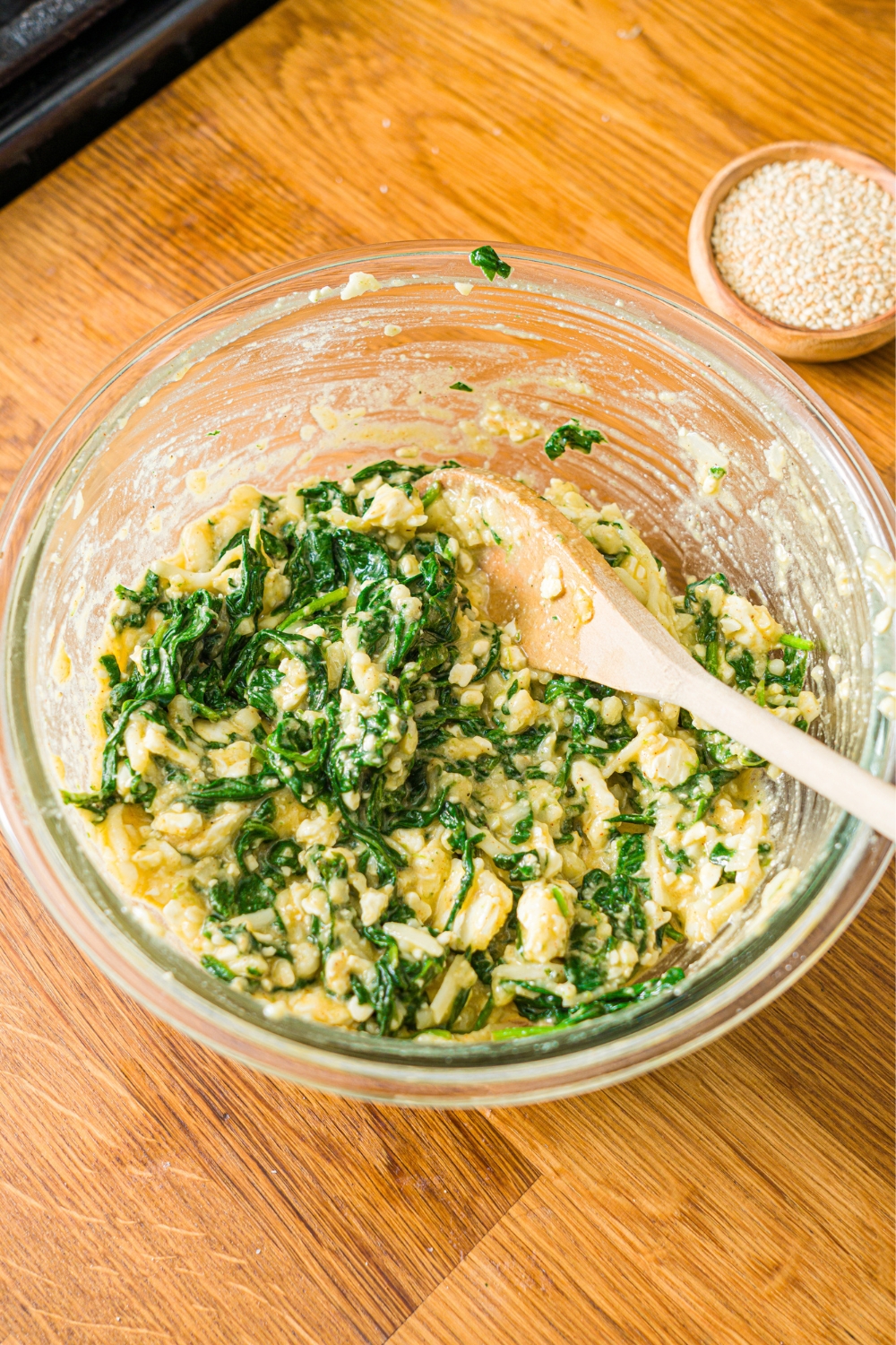 A glass bowl with a wooden spoon stirring spanakopita filling including spinach, egg, cheese, and seasonings. The bowl is on a wooden counter.