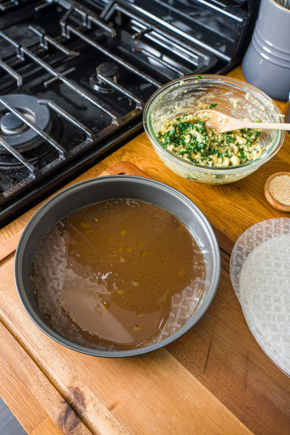 A tin with rice paper sheets soaking in an egg and soy sauce mixture. The tin is on a wooden counter with a bowl of spanakopita filling.
