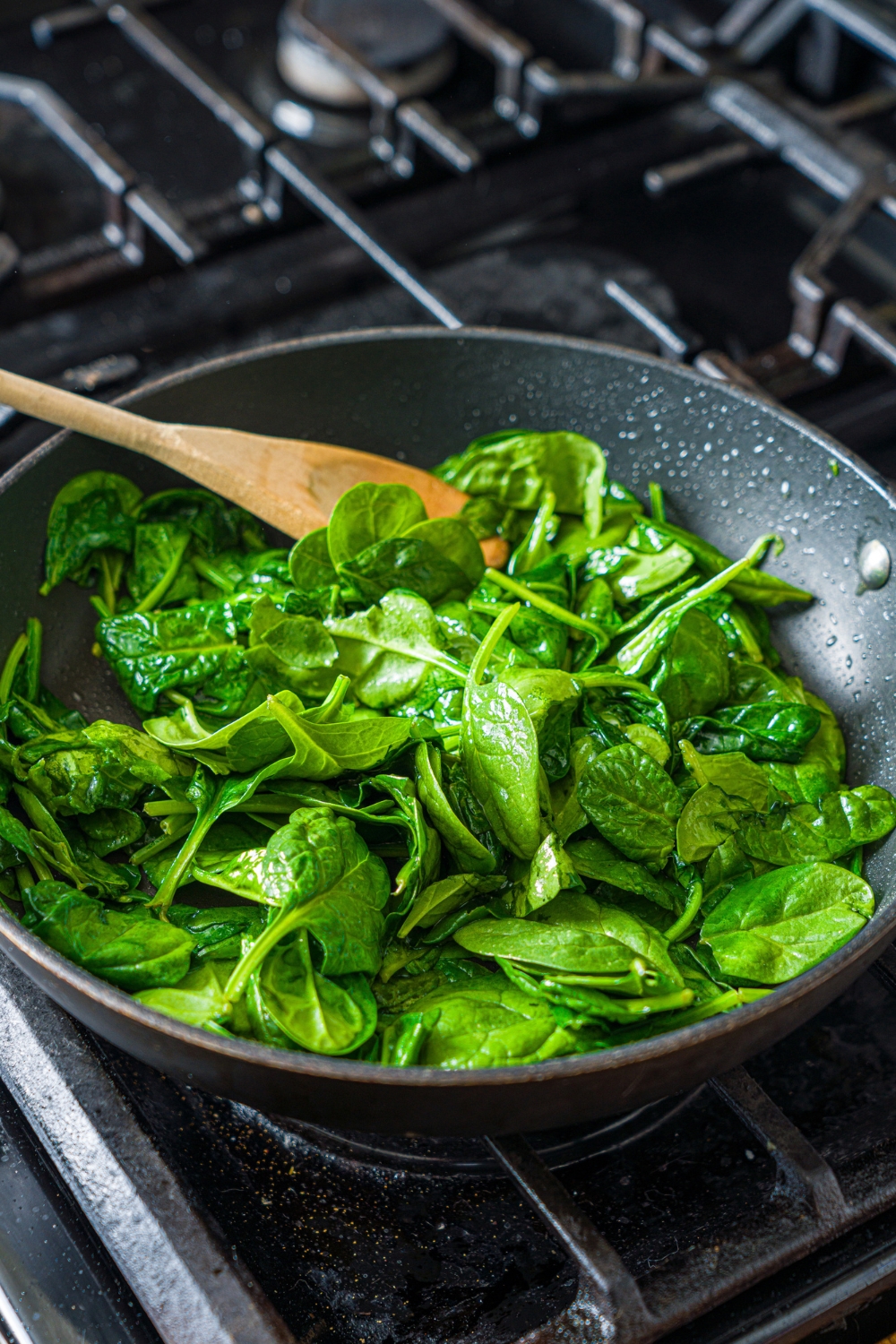An oven with a skillet cooking spinach with a wooden spoon.