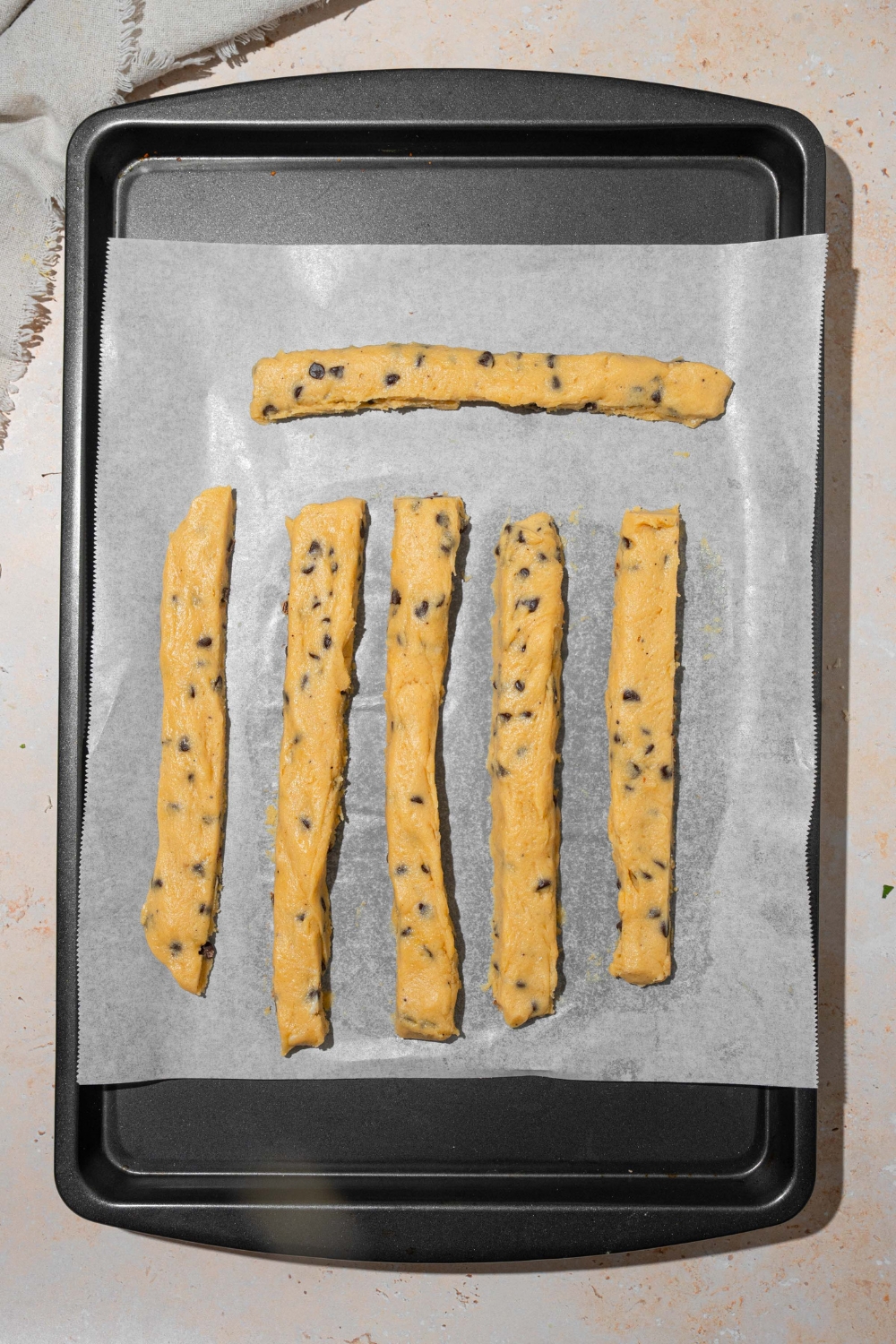A baking sheet lined with parchment paper with uncooked chocolate chip cookie fries. The sheet is on a tan counter.