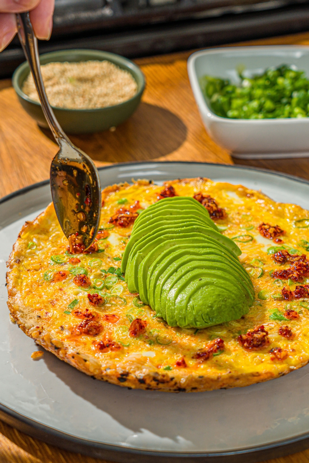 A white plate with a crispy rice omelette topped with sliced avocado. A spoon is pouring chili crunch oil over the omelette. The plate is on a wooden counter with bowls of garnishes.