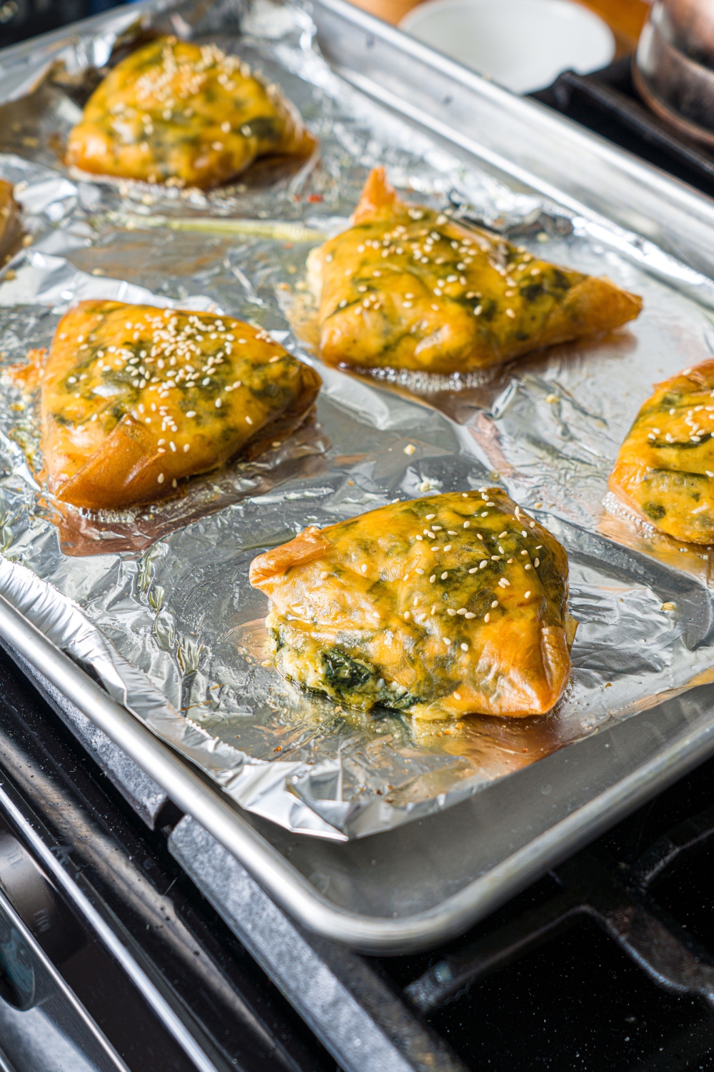 A baking sheet lined with foil with several pieces of baked rice paper spanakopita topped with sesame seeds.