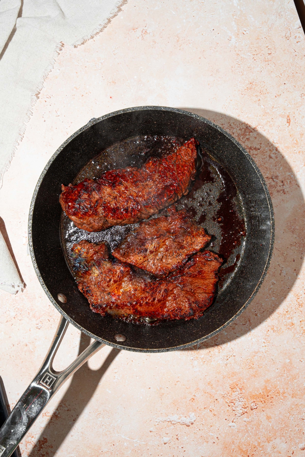Three marinated skirt steaks cooking in a skillet. The skillet is on a tan counter.