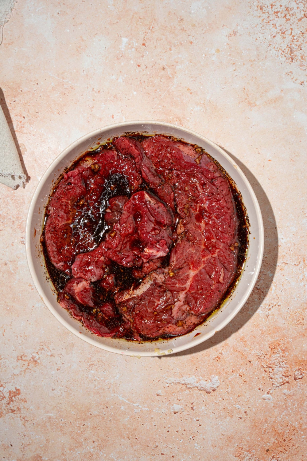 A white bowl with skirt steaks marinating in a soy sauce and balsamic vinegar mixture. The plate is on a tan counter.