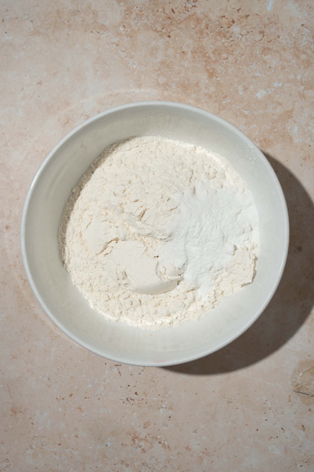 A white bowl with ingredients to make cookie dough including flour, baking powder, baking soda, and salt. The bowl is on a tan counter.