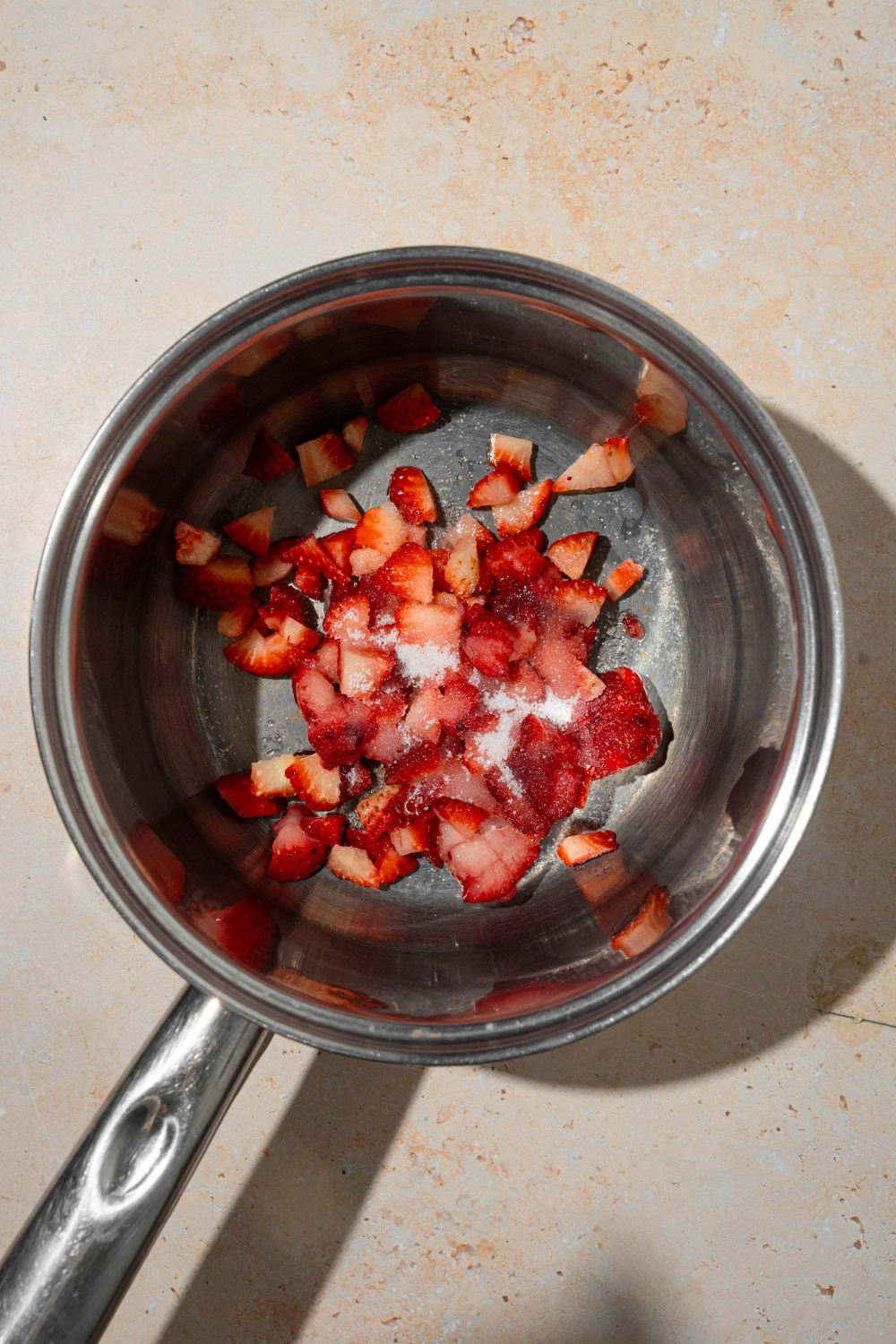 A stock pot with diced strawberries and sugar. The pot is on a tan counter.