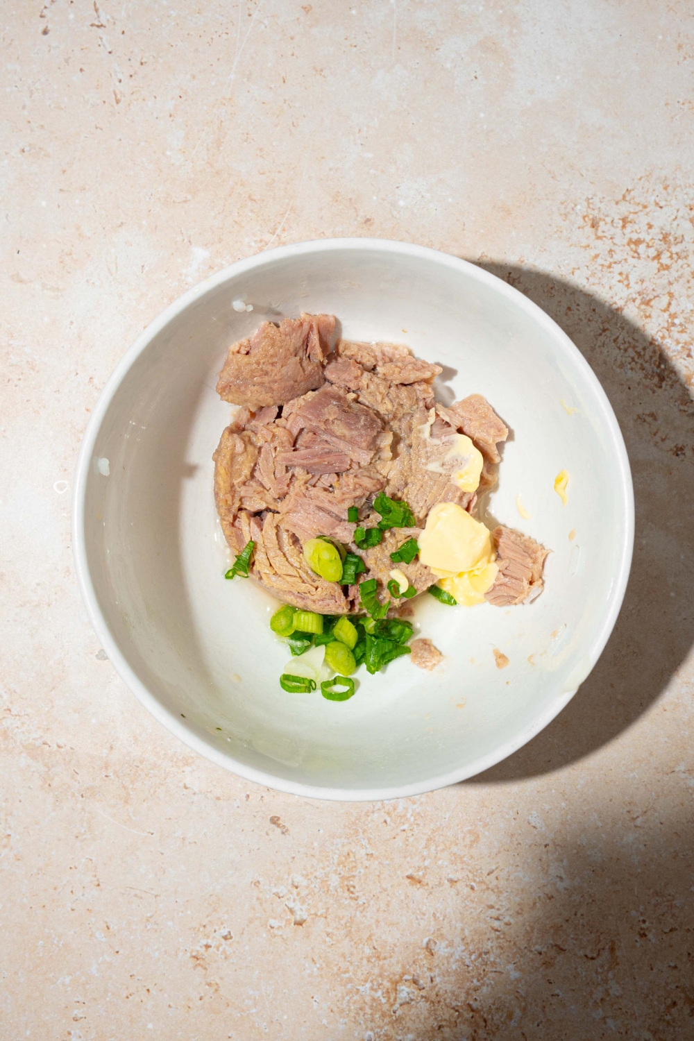 A white bowl with ingredients for tuna salad including canned tuna, sliced green onion, and mayo. The bowl is on a tan counter.