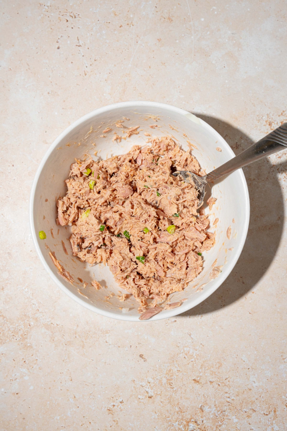 A bowl with a fork mixing tuna salad. The bowl is on a tan counter.