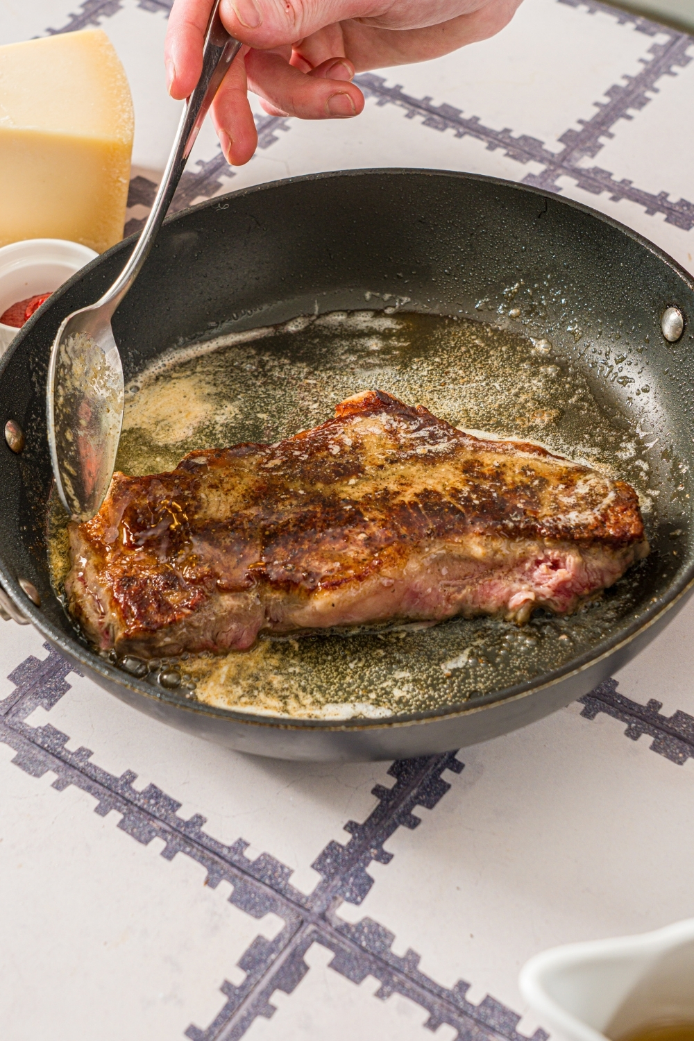 A skillet with a browned steak cooking in butter and oil. A spoon is basting the steak. The skillet is on a tiled counter.