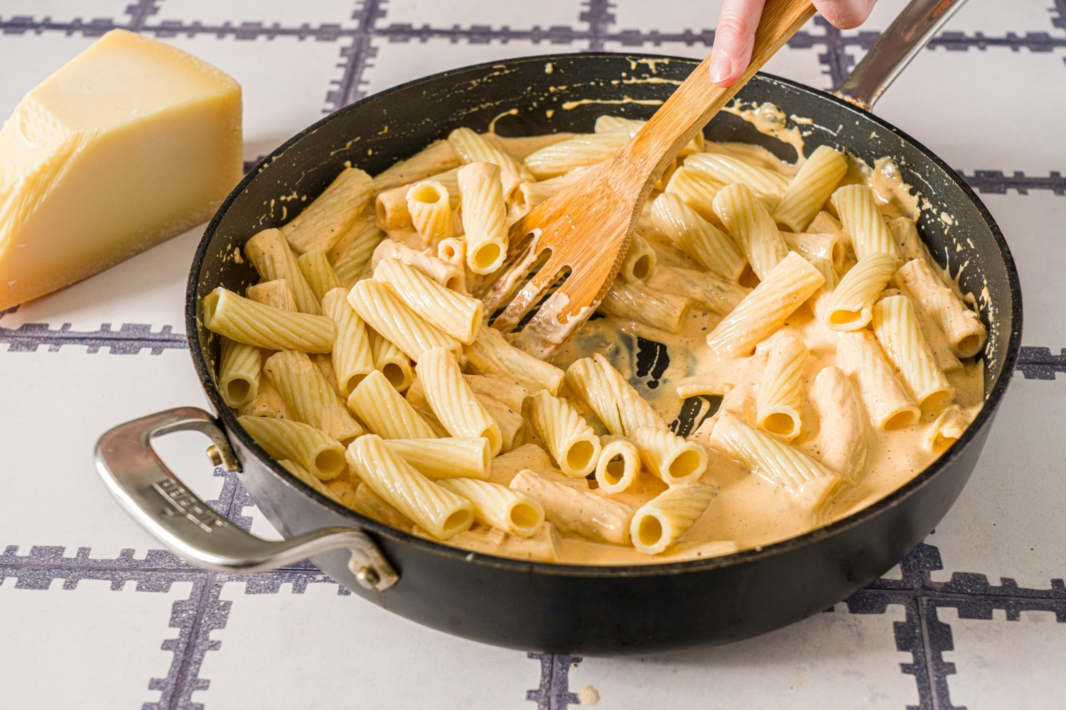A skillet with a wooden spoon mixing rigatoni pasta with a cajun cream sauce. The skillet is on a tiled counter.