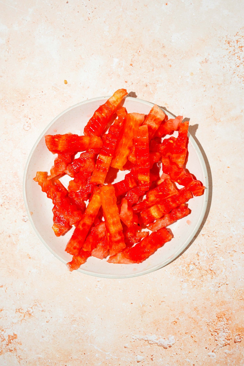 A white plate with watermelon sliced into fry shapes. The plate is on a tan counter.