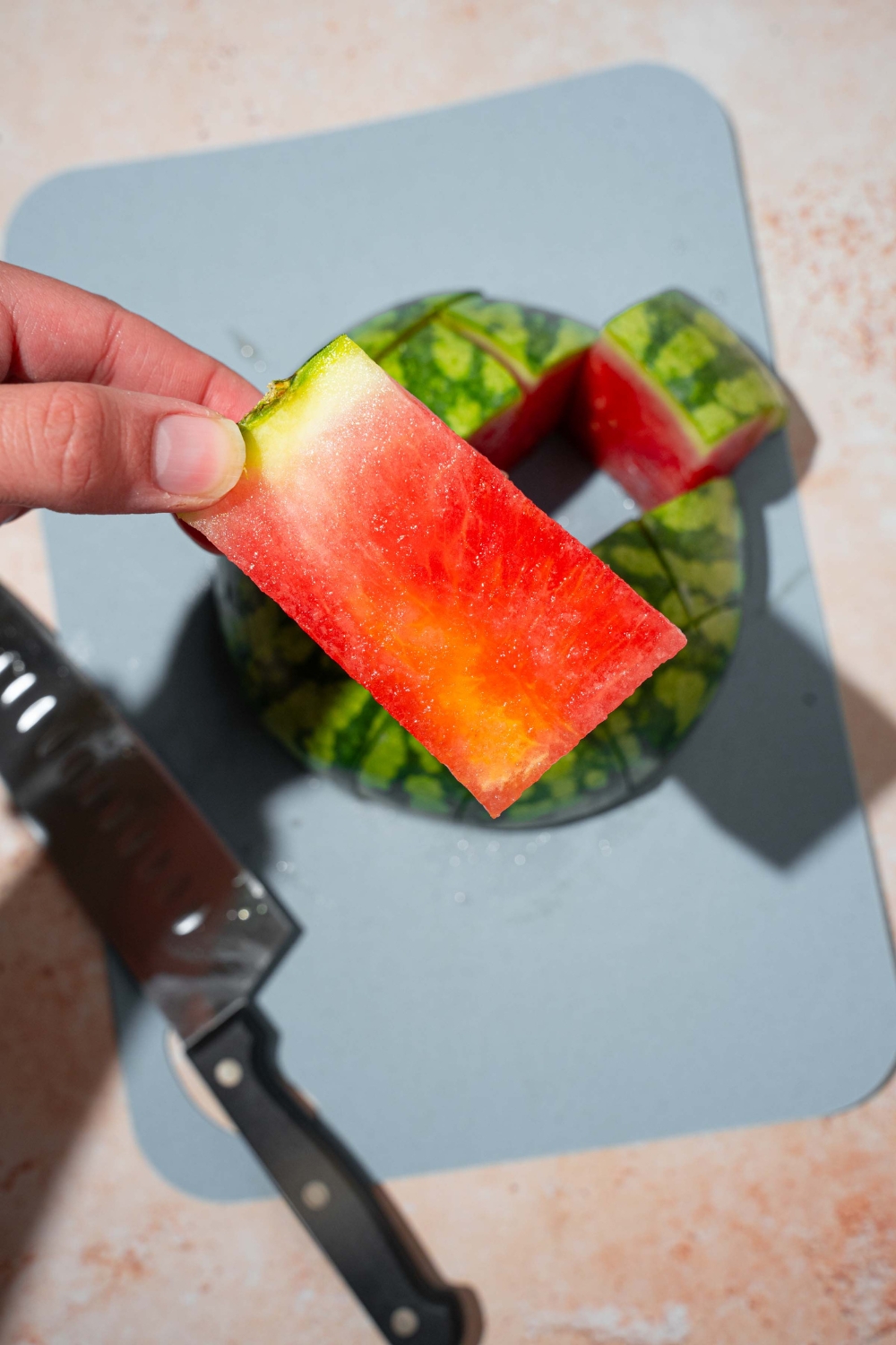 A hand holding a frozen watermelon wedge. There is a blue cutting board with frozen watermelon and a knife.