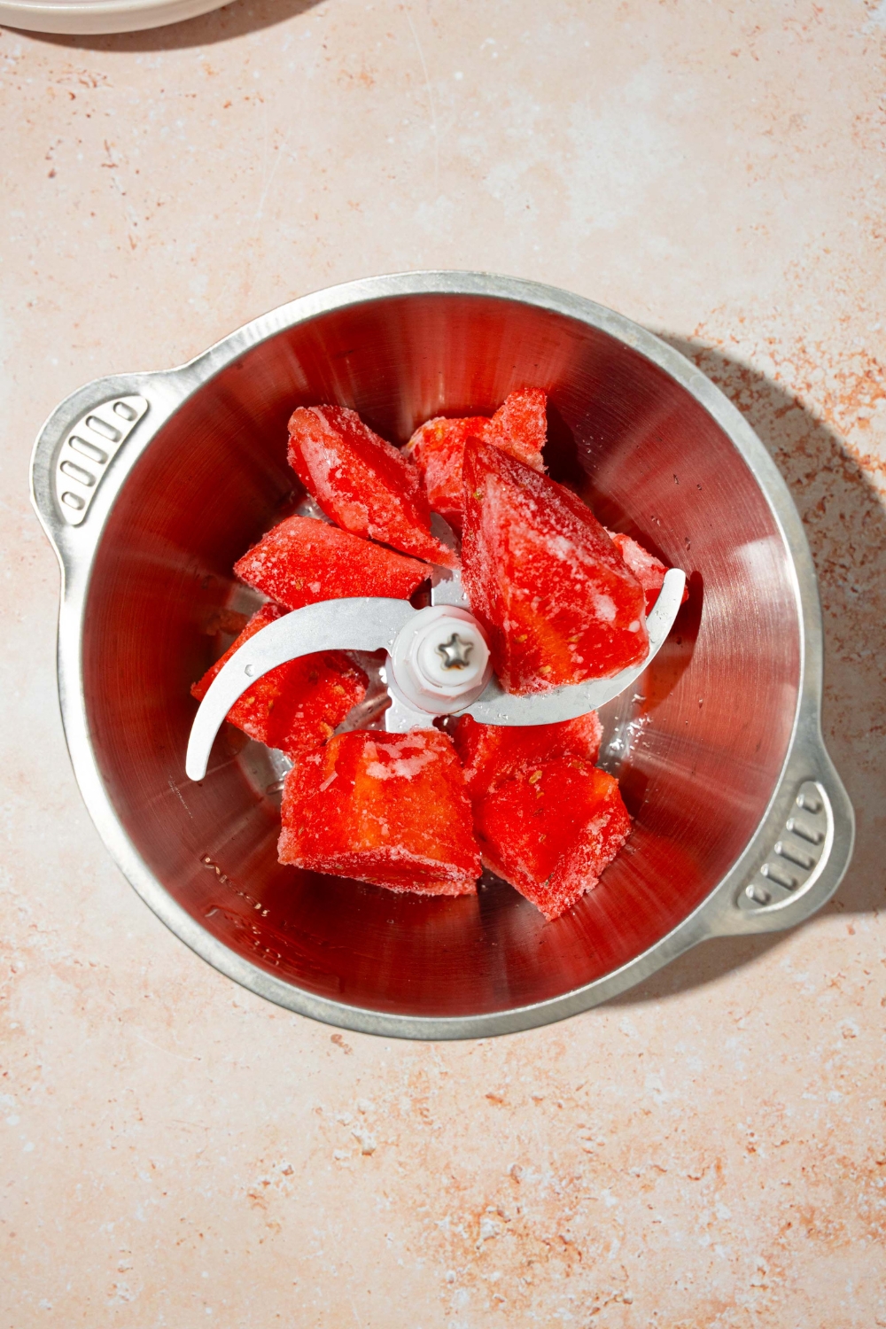 A food processor bowl with frozen chunks of watermelon. The bowl is on a tan counter.