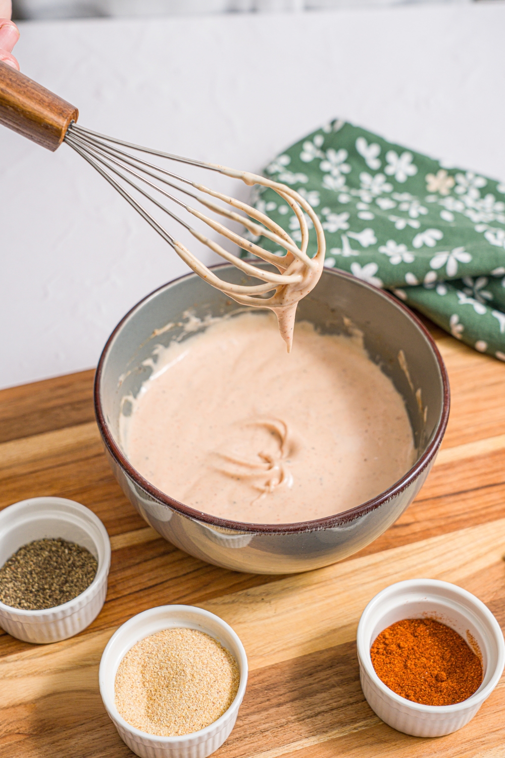 A bowl of mixed yum yum hibachi sauce. A whisk is being dipped into the bowl. The bowl is on a wooden board with small bowls of seasonings. The board is on a white counter with a green floral napkin.