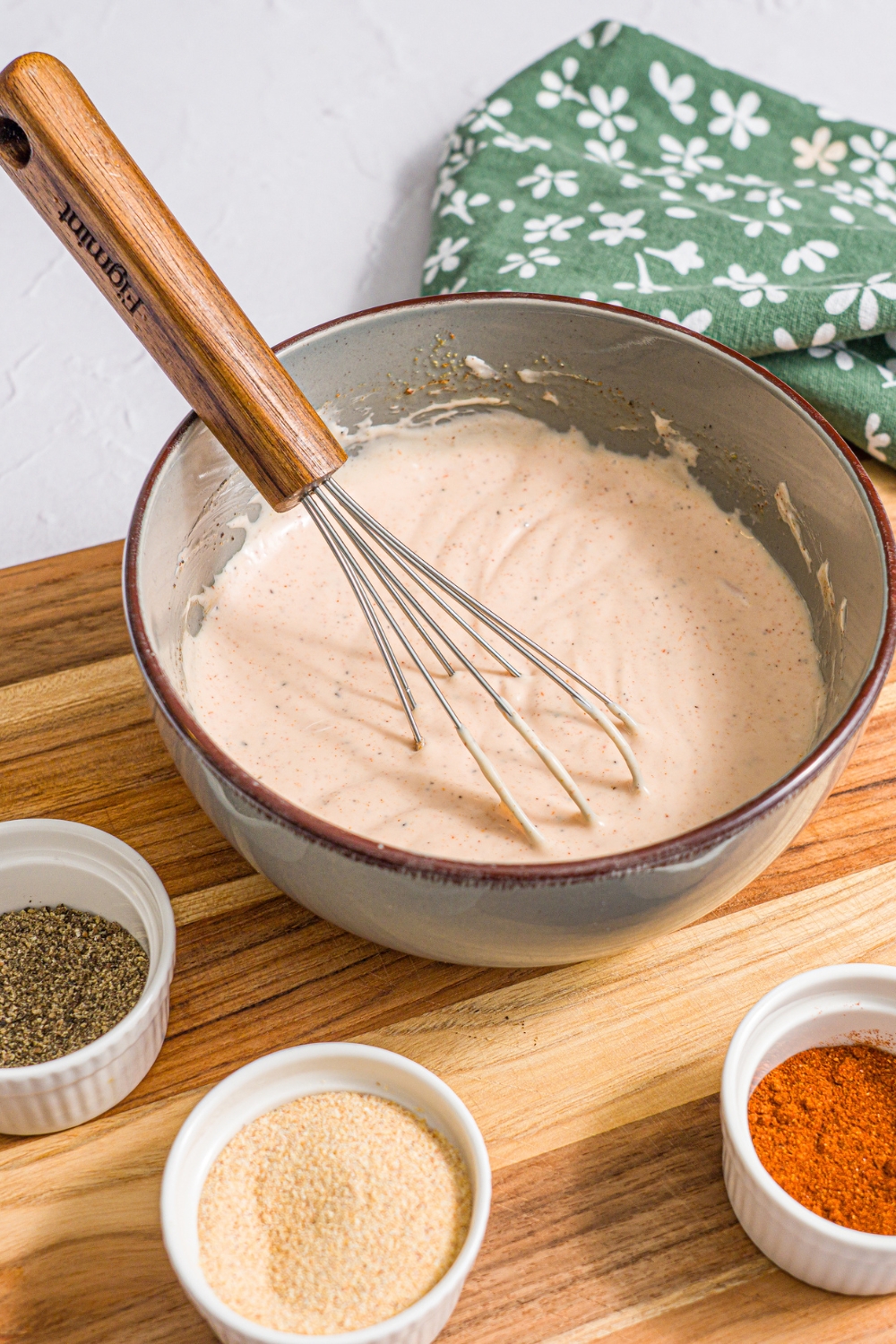 A bowl of mixed yum yum hibachi sauce. A whisk is in the bowl. The bowl is on a wooden board with small bowls of seasonings. The board is on a white counter with a green floral napkin.