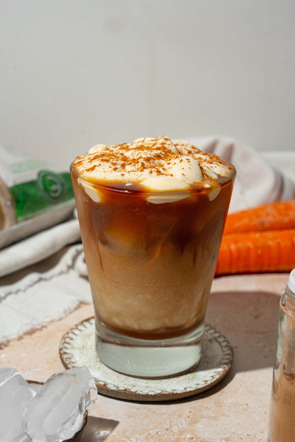 A glass tumbler with iced carrot cake latte topped with vanilla cream cheese foam and sprinkled with cinnamon. The glass is on a ceramic coaster on a tan counter with a white cloth napkin and carrots.
