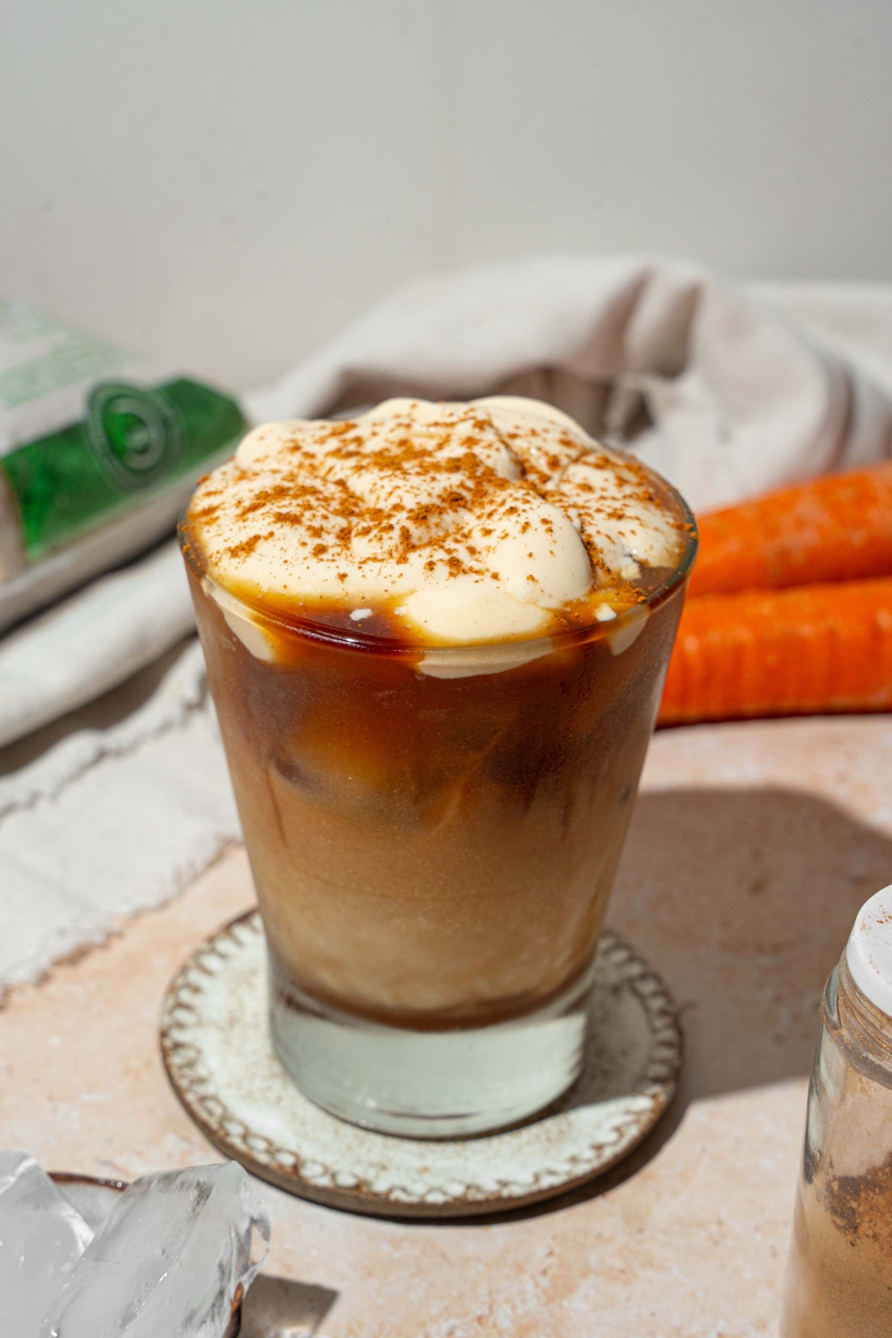 A glass tumbler with iced carrot cake latte topped with vanilla cream cheese foam and sprinkled with cinnamon. The glass is on a ceramic coaster on a tan counter with a white cloth napkin and carrots.