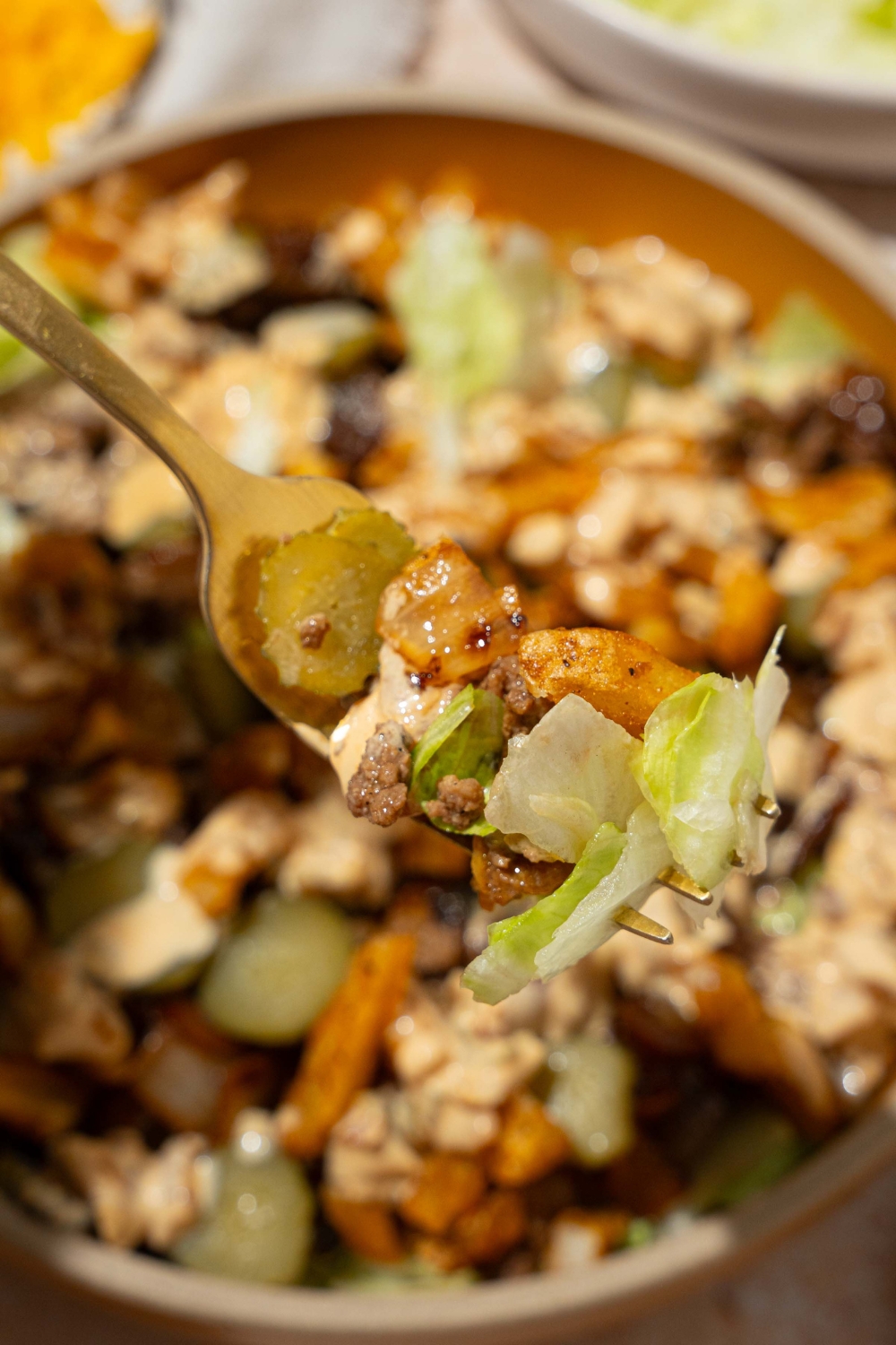 A fork with a bite of In-n-Out burger bowl with shredded lettuce, seasoned burger meat, and french fries. There is a burger bowl blurred in the background.