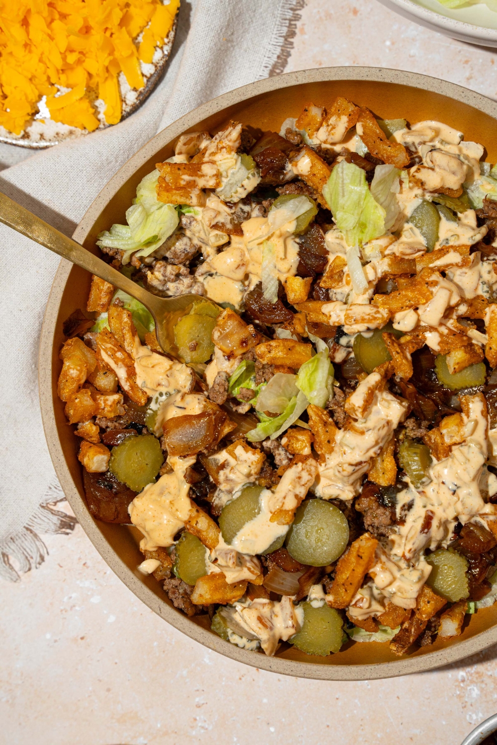 An orange bowl with an In-n-Out burger bowl with shredded lettuce, seasoned burger meat, french fries, pickles, and In-n-Out sauce. A fork is in the plate. The plate is on a tan counter with a small plate of shredded cheese.
