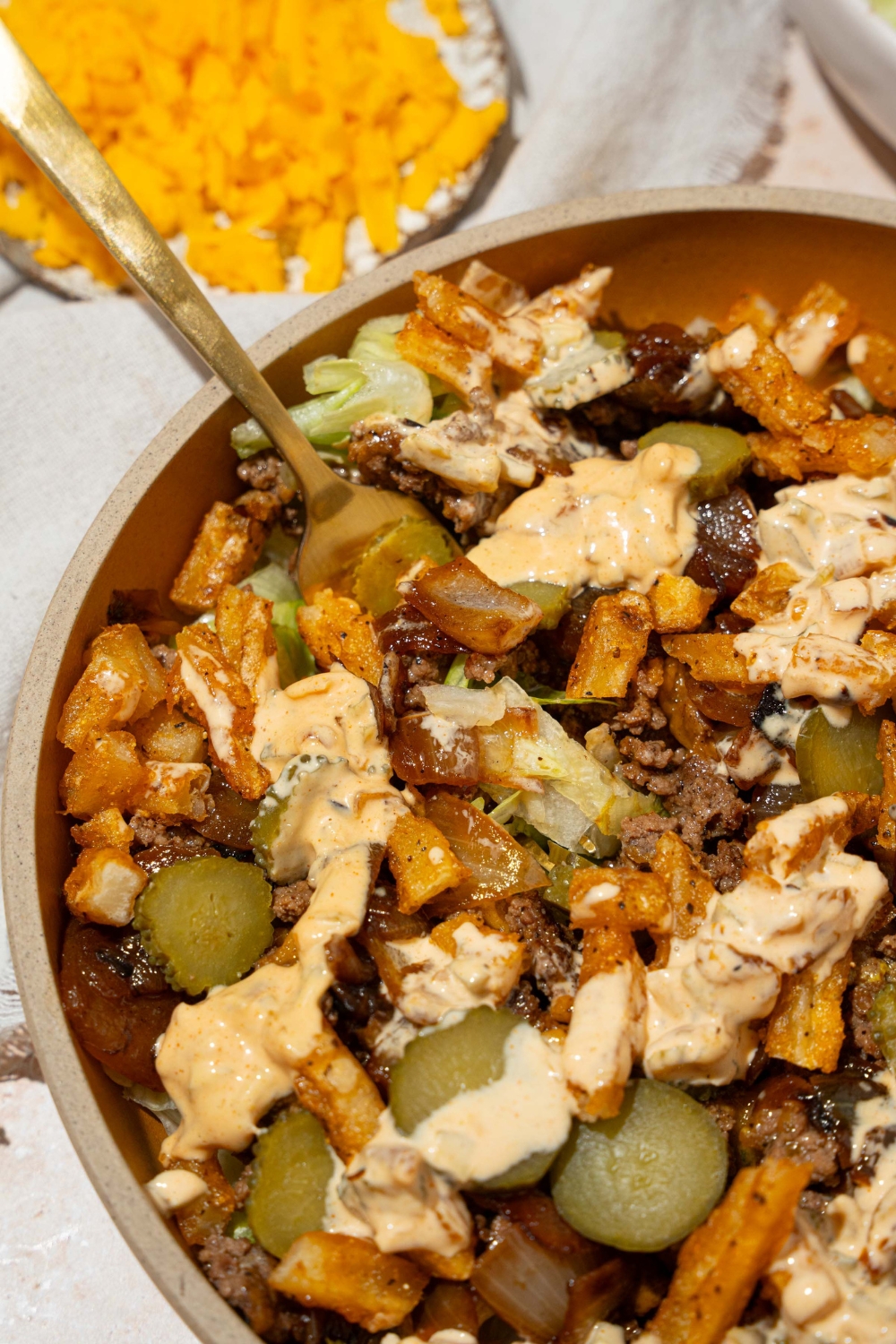 An orange bowl with an In-n-Out burger bowl with shredded lettuce, seasoned burger meat, french fries, pickles, and In-n-Out sauce. A fork is in the plate. The plate is on a tan counter with a small plate of shredded cheese.