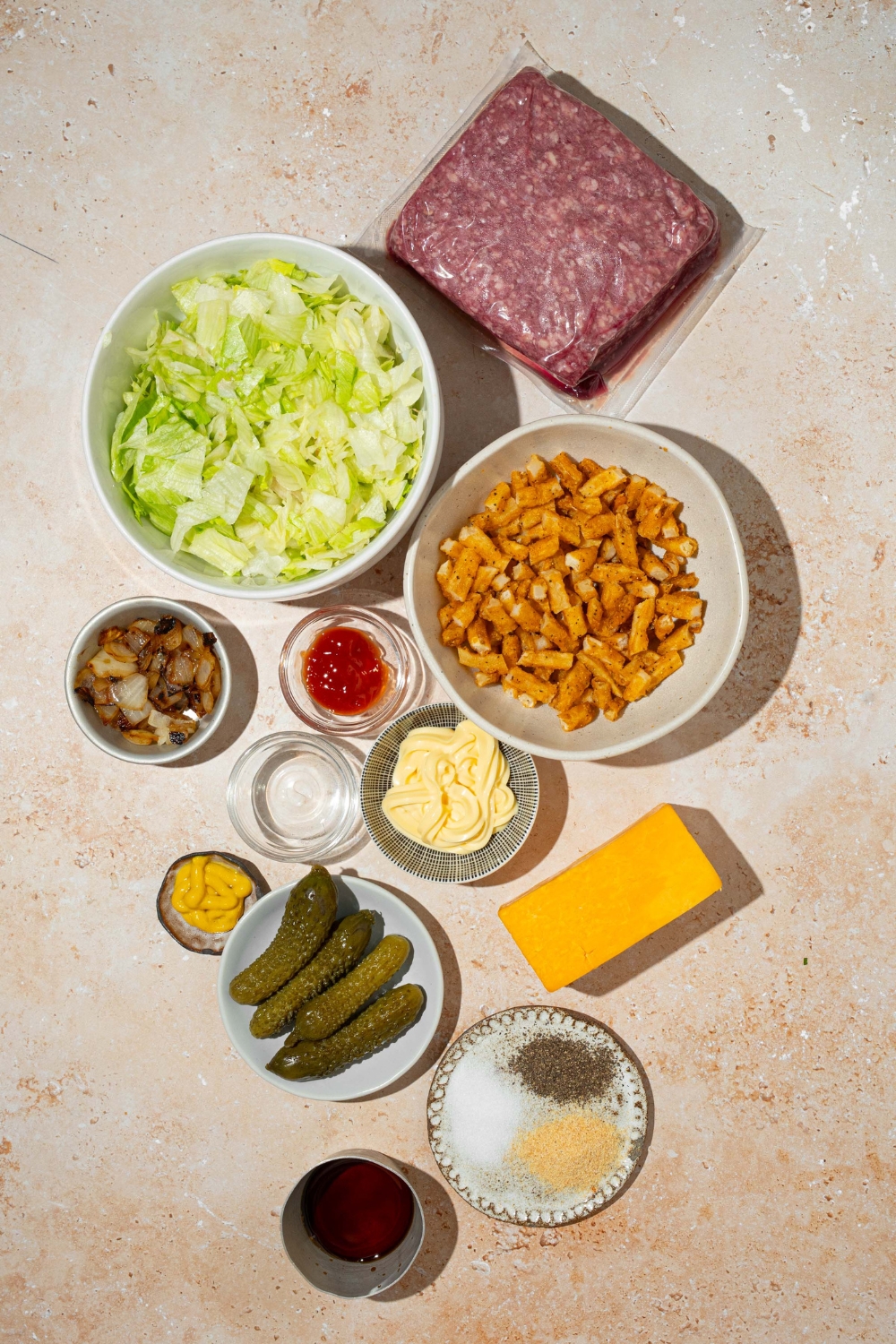 An overhead shot of several bowls in various sizes containing ingredients to make In-n-Out burger bowls including ground beef, shredded lettuce, french fries, pickles, caramelized onion, ketchup, mayo, mustard, cheddar cheese, and seasonings.
