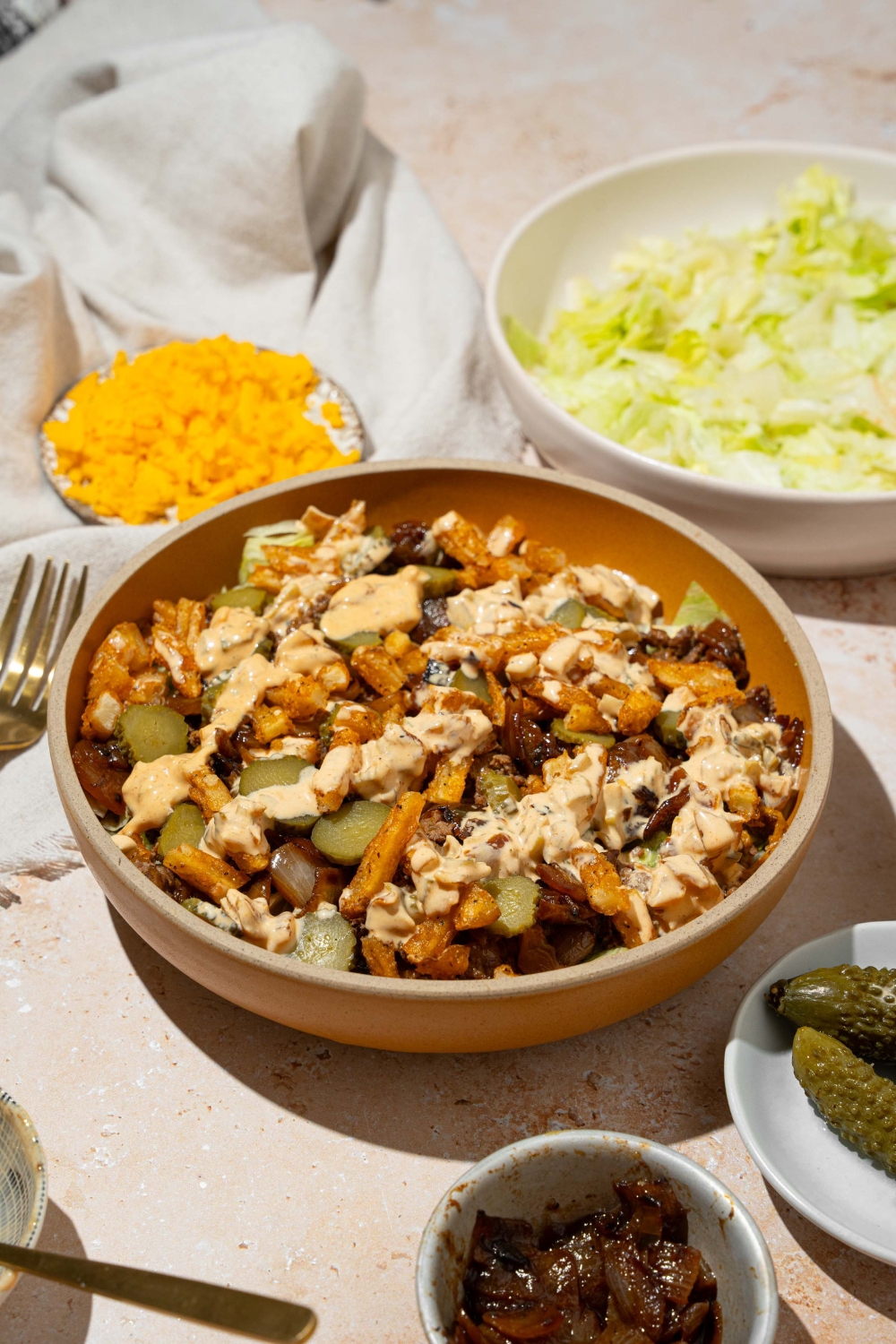 An orange bowl with an In-n-Out burger bowl with shredded lettuce, seasoned burger meat, french fries, pickles, and In-n-Out sauce. The plate is on a tan counter with plates of ingredients and a white cloth napkin.