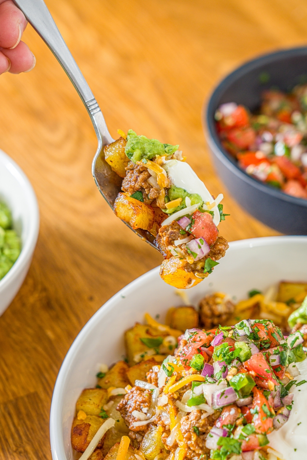 A fork with a bite of loaded potato taco bowl over a white bowl including roasted potatoes, taco meat, shredded cheese, pico de gallo, sour cream, and guacamole. The bowl is garnished with cilantro.