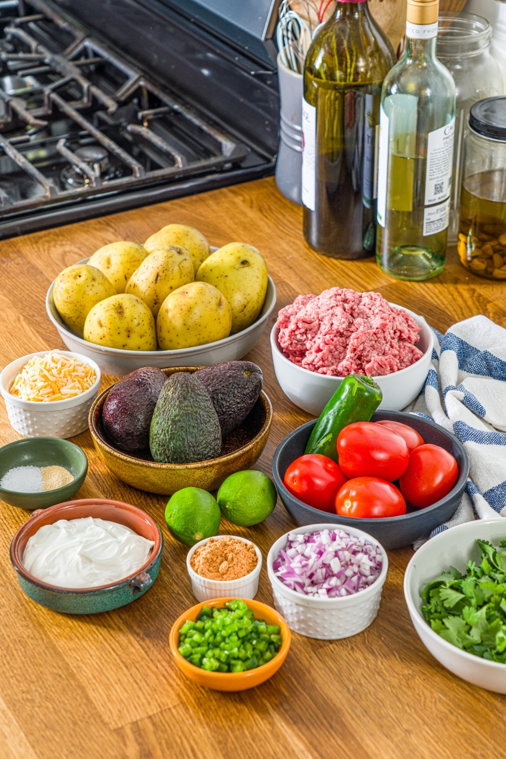 A wooden counter with several bowls in various sizes containing ingredients to make loaded potato taco bowl including avocado, potatoes, ground beef, tomatoes, lime, shredded cheese, sour cream, cilantro, and seasonings.