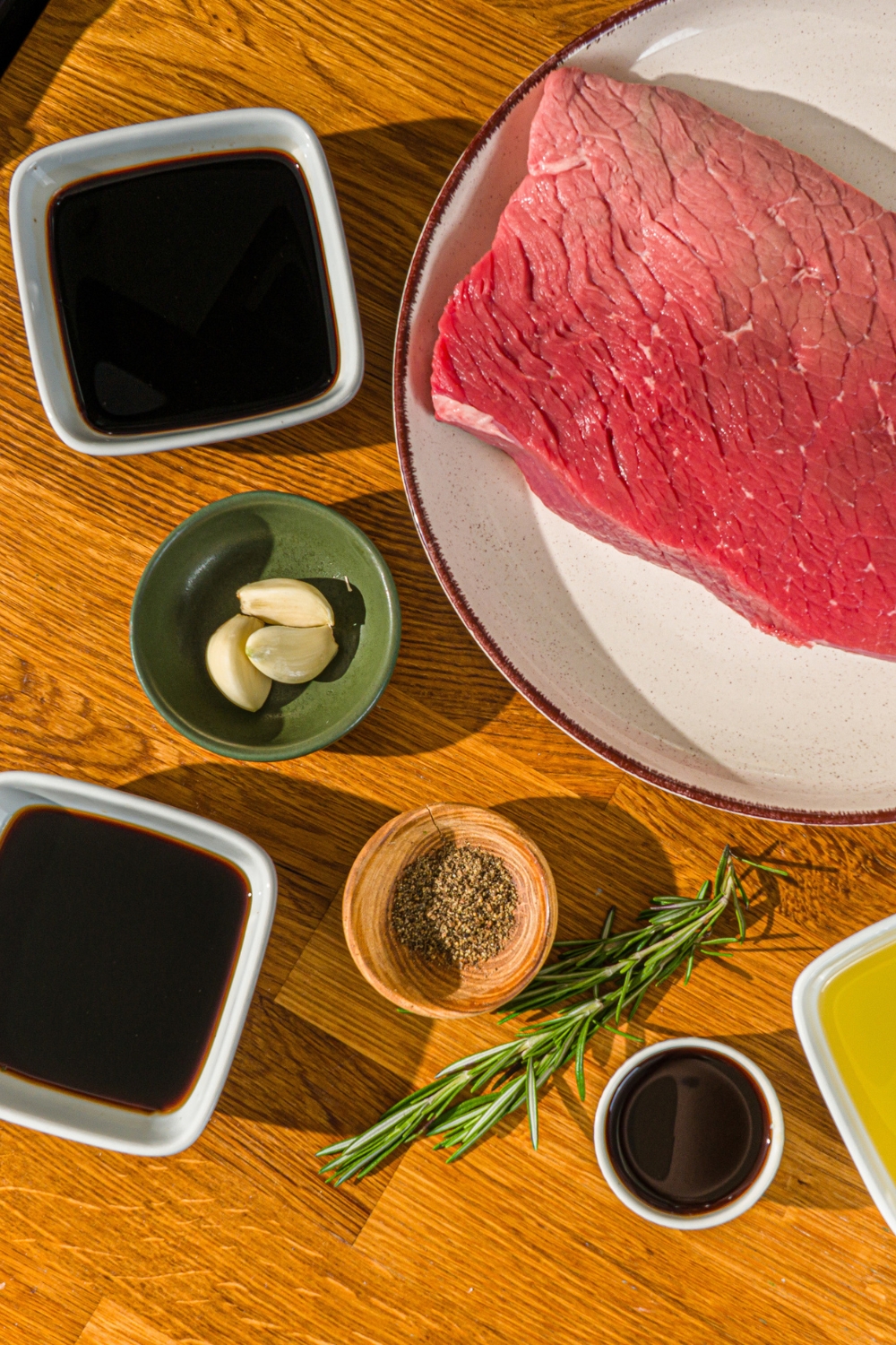 A wooden counter with several bowls in various sizes containing ingredients to make London broil marinade including London broil beef, soy sauce, balsamic vinegar, red wine vinegar, Worcestershire sauce, garlic, rosemary, and black pepper.
