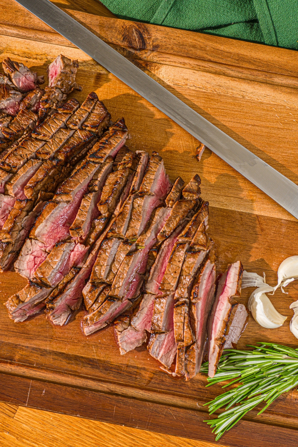 A wooden board with sliced London broil served with garlic and rosemary. There is a knife on the board.