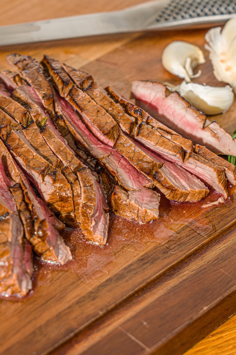 A wooden board with sliced London broil served with garlic and rosemary. There is a knife on the board.