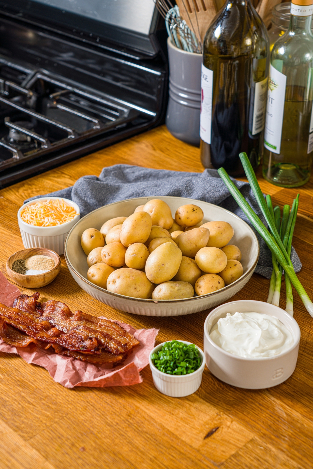 A wooden counter with several bowls in various sizes containing ingredients to make mini baked potatoes including mini potatoes, bacon, sour cream, shredded cheese, green onion, and seasonings.