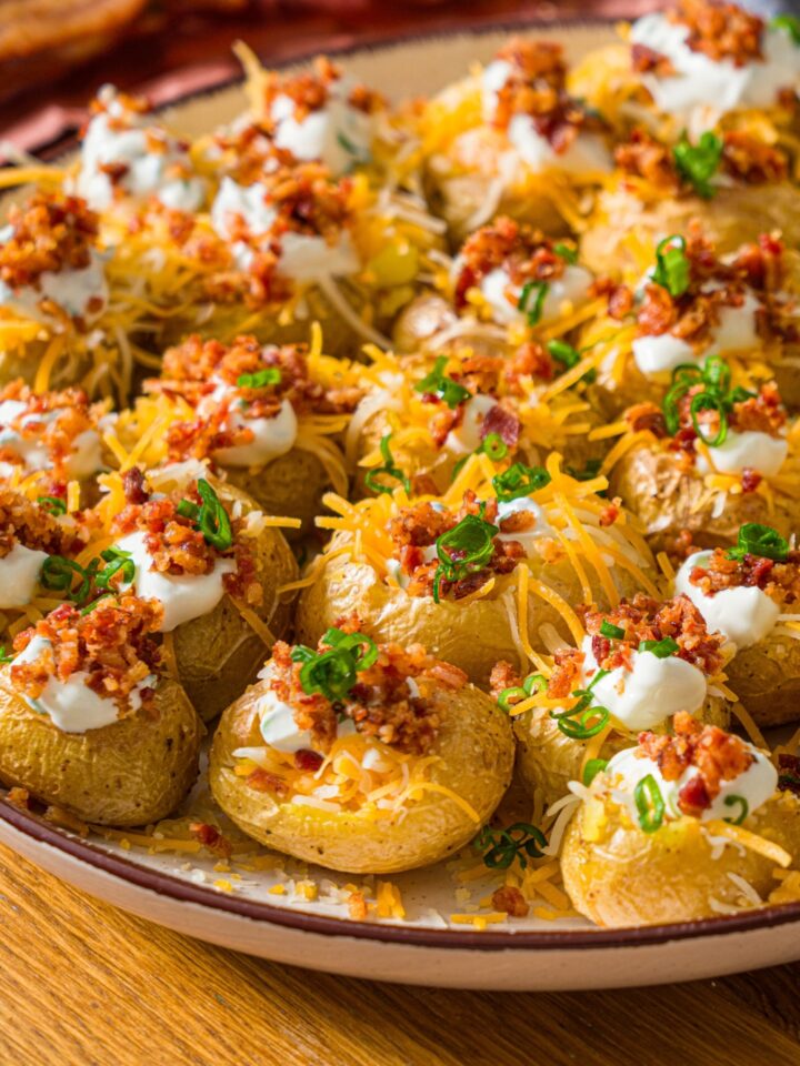 A white plate with mini baked potatoes loaded with sour cream, shredded cheese, and crumbled bacon, garnished with sliced green onion.