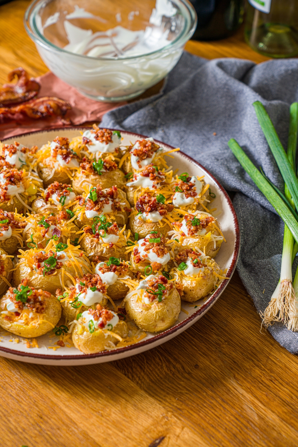 A white plate with mini baked potatoes loaded with sour cream, shredded cheese, and crumbled bacon, garnished with sliced green onion. The plate is on a wooden board with a blue cloth napkin and green onions.