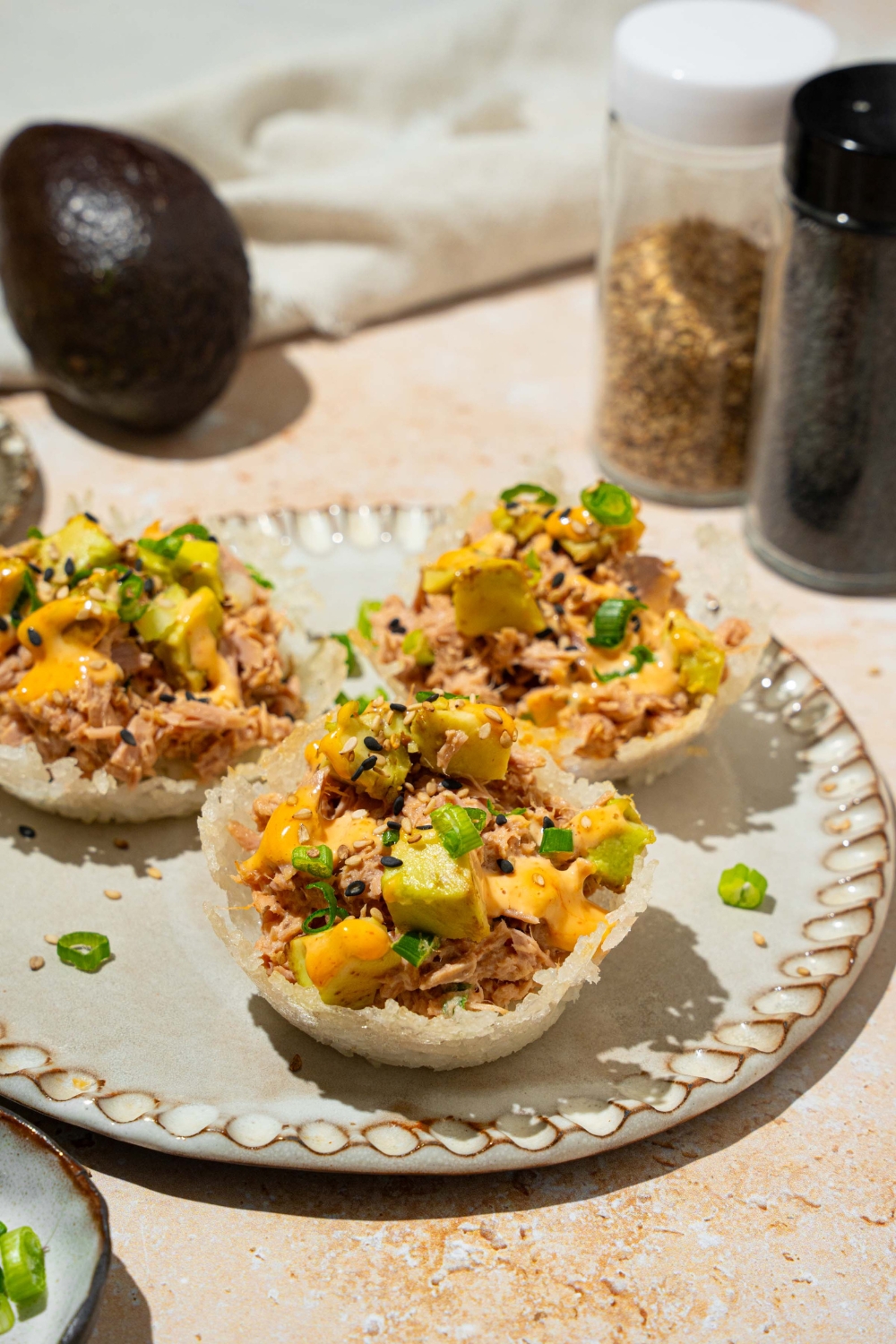 A plate with three tuna rice cups topped with avocado, sliced green onion, sesame seeds, and spicy mayo. The plate is on a tan counter with a white cloth napkin and avocado.