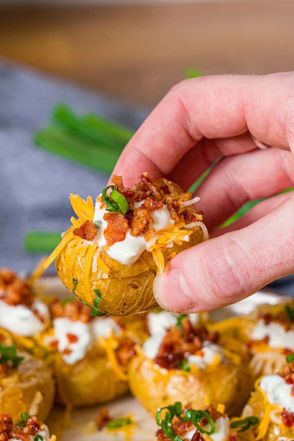 A hand holding a mini baked potato topped with sour cream, shredded cheese, crumbled bacon, and garnished with sliced green onion. There is a plate of mini potatoes blurred in the background.