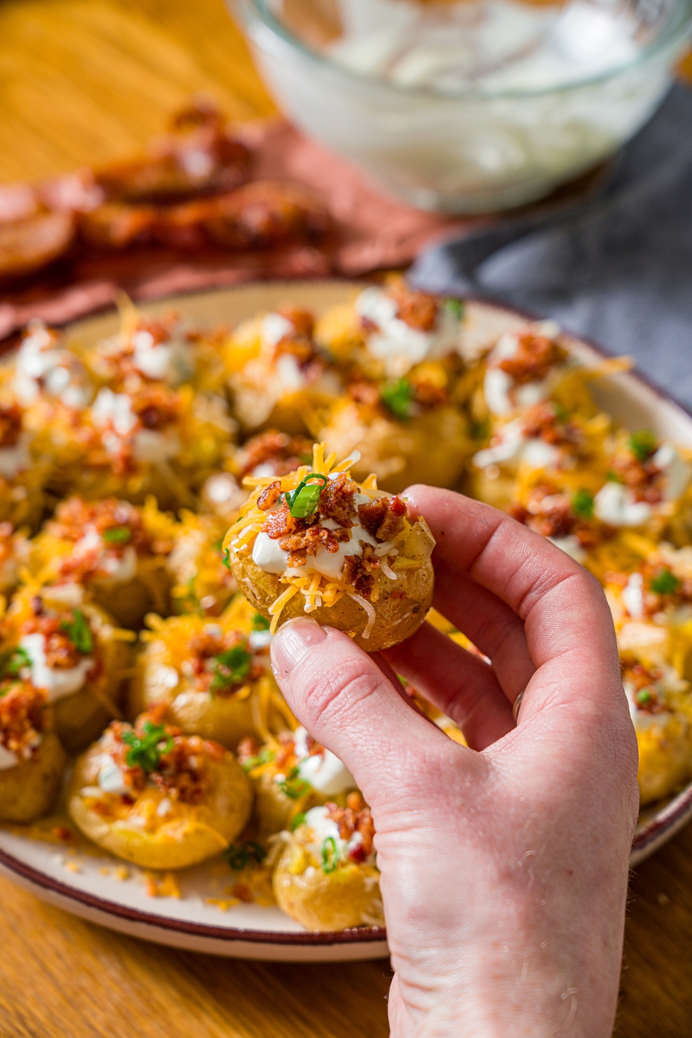 A hand holding a mini baked potato topped with sour cream, shredded cheese, crumbled bacon, and garnished with sliced green onion. There is a plate of mini potatoes blurred in the background.