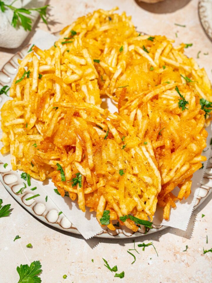A plate lined with parchment paper with crispy parmesan hashbrowns garnished with fresh parsley. The plate is on a tan counter.
