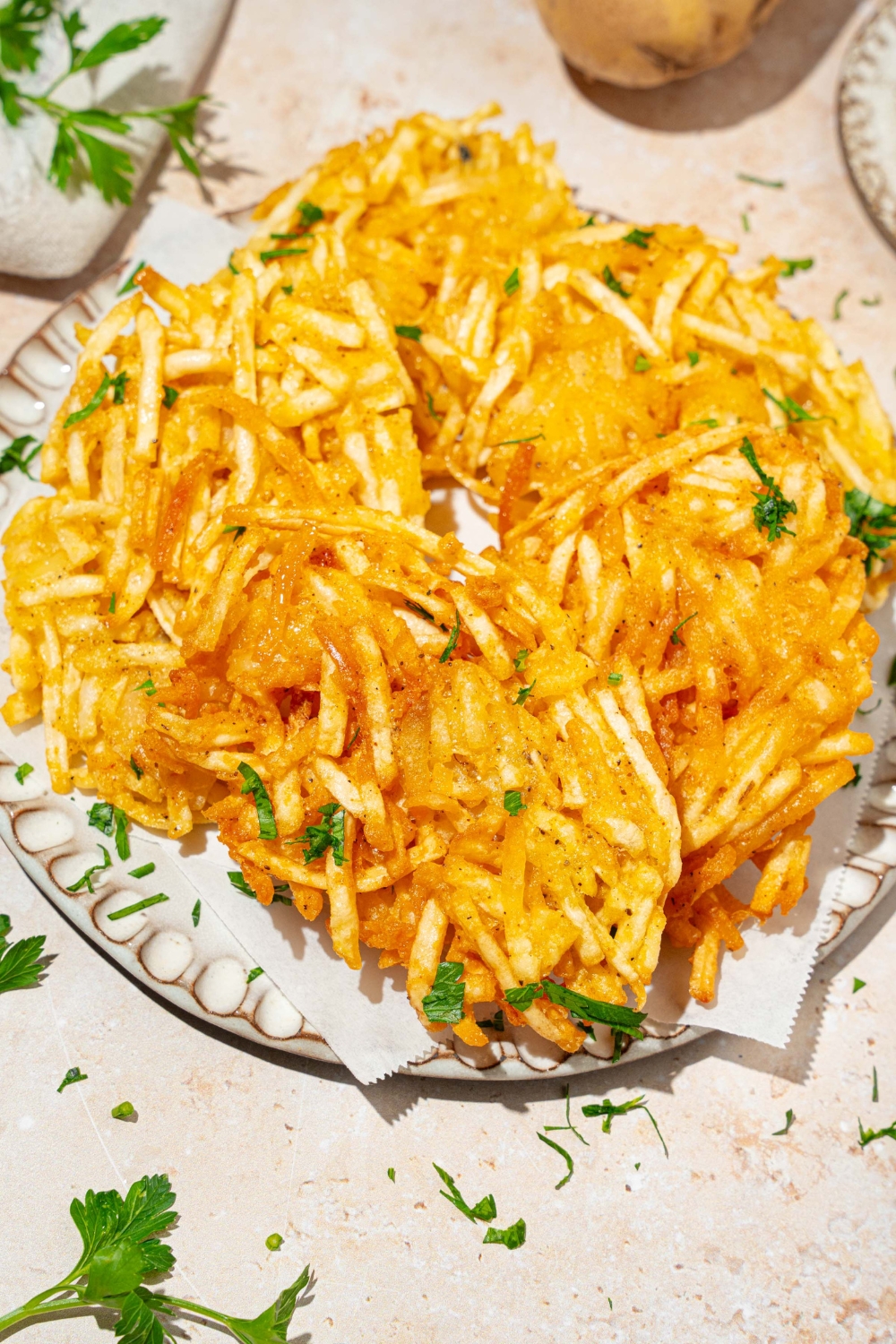 A plate lined with parchment paper with crispy parmesan hashbrowns garnished with fresh parsley. The plate is on a tan counter.