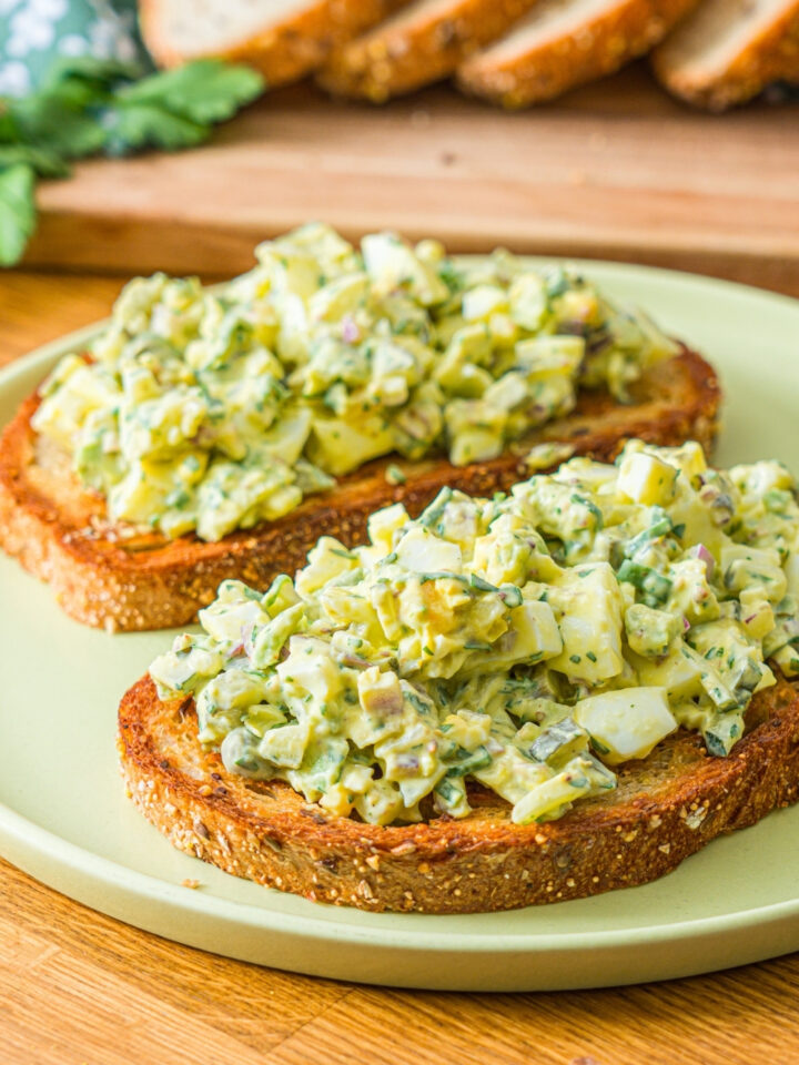 A green plate with two slices of toasted bread topped with pickle egg salad garnished with sliced green onion and served with a pickle spear. The plate is on a wooden counter with a wooden board of sliced bread with garnishes.