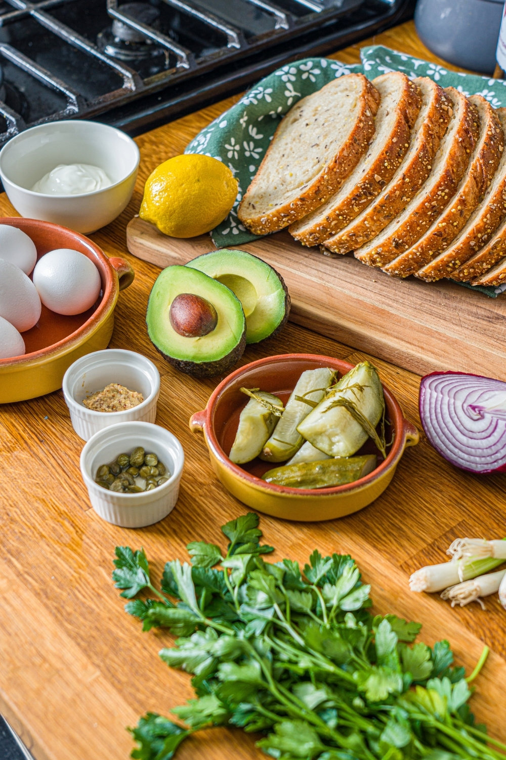 A wooden board with ingredients to make pickle egg salad including eggs, pickles, avocado, capers, red onion, parsley, greek yogurt, and sliced bread.