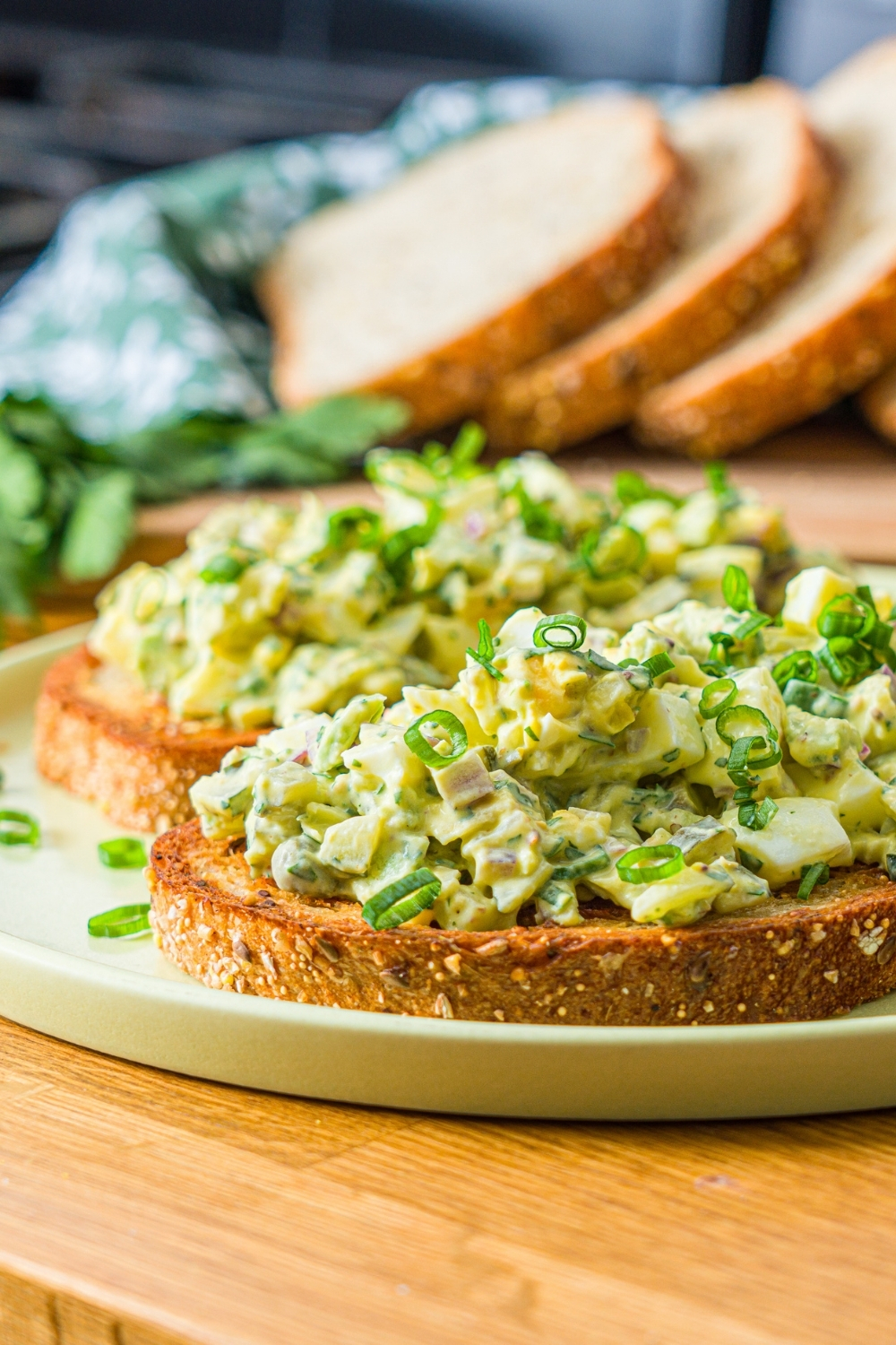 A green plate with two slices of toasted bread topped with pickle egg salad garnished with sliced green onion and served with a pickle spear. The plate is on a wooden counter with a wooden board of sliced bread with garnishes.