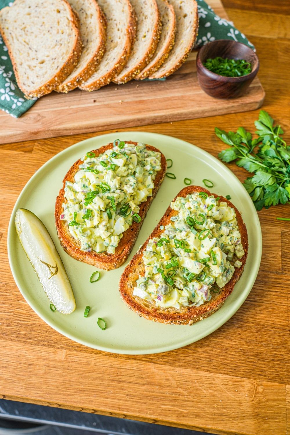 A green plate with two slices of toasted bread topped with pickle egg salad garnished with sliced green onion and served with a pickle spear. The plate is on a wooden counter with a wooden board of sliced bread with garnishes.