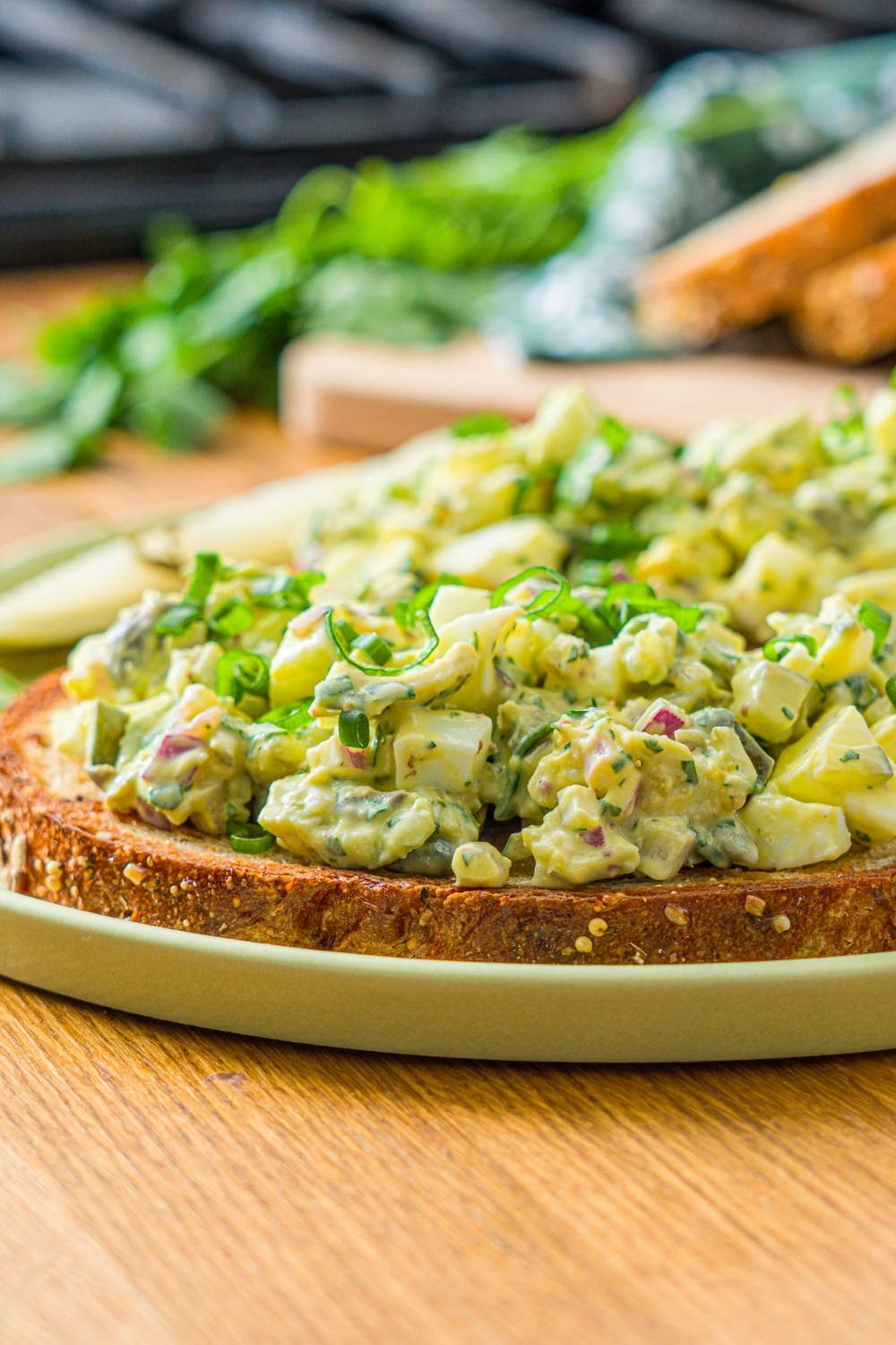 A green plate with two slices of toasted bread topped with pickle egg salad garnished with sliced green onion and served with a pickle spear. The plate is on a wooden counter with a wooden board of sliced bread with garnishes.