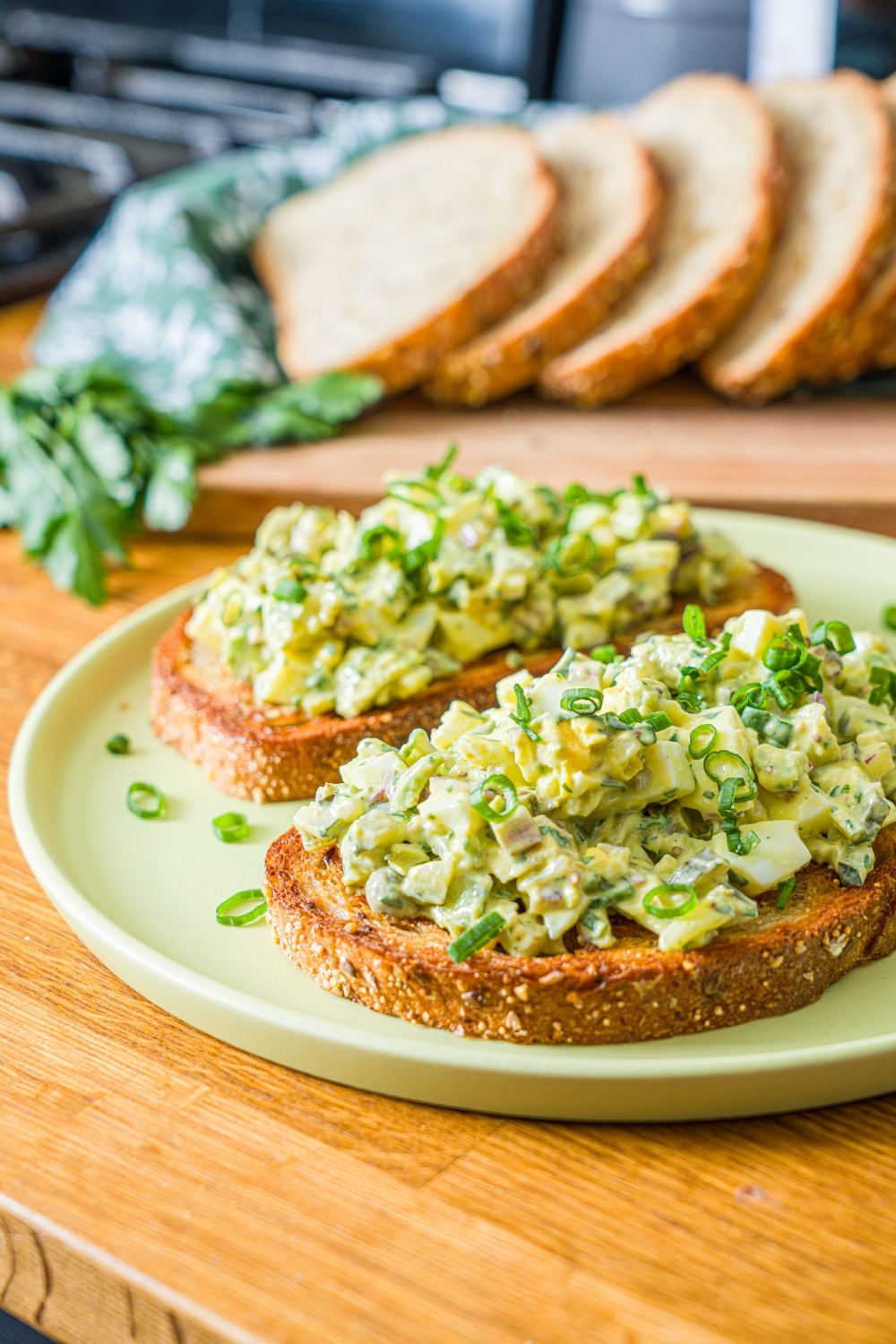 A green plate with two slices of toasted bread topped with pickle egg salad garnished with sliced green onion and served with a pickle spear. The plate is on a wooden counter with a wooden board of sliced bread with garnishes.