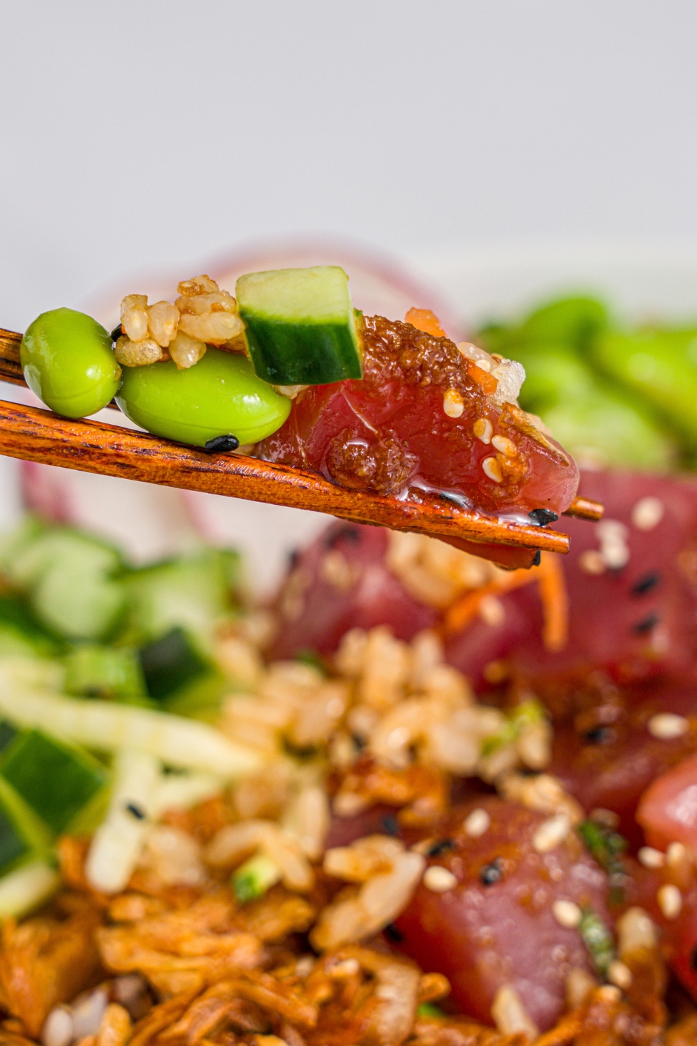 A close up of a pair of chopsticks with tuna poke, edamame, and rice. There is a large tuna poke bowl garnished with sesame seeds and crispy onions blurred in the background.