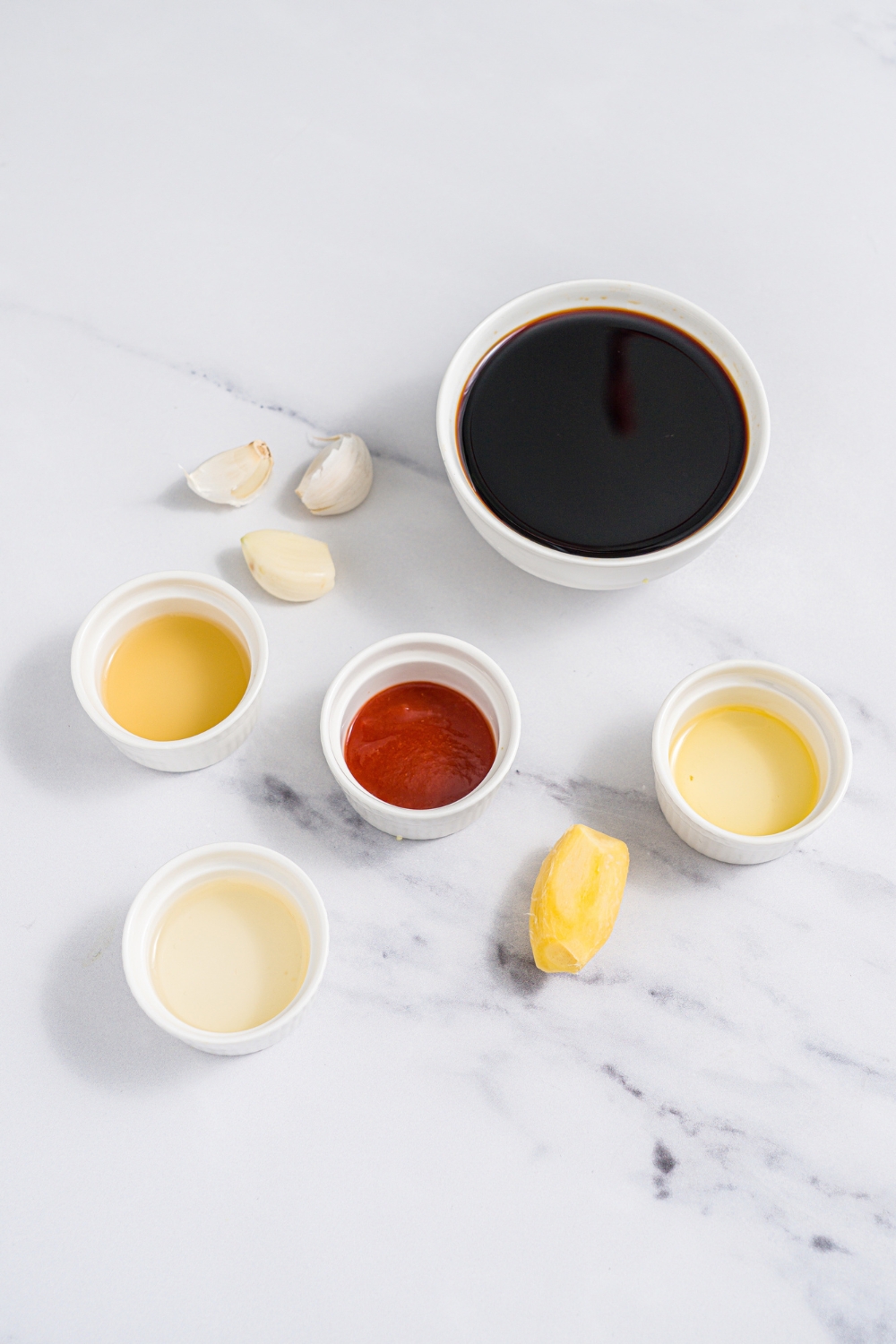 A marble counter with several bowls in various sizes containing ingredients to make poke sauce including soy sauce, rice vinegar, sesame oil, garlic, ginger, lime juice, and sriracha