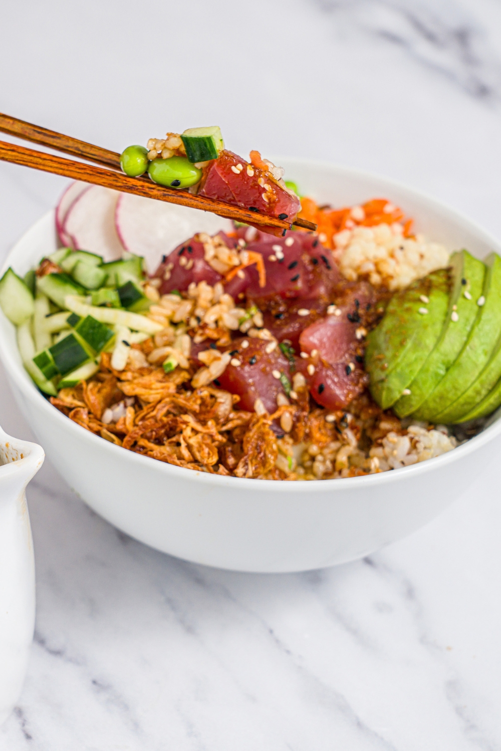 A tuna poke bowl in a white bowl with rice, avocado, edamame, crispy onions, cucumber radishes, and sesame seeds. A pair of chopsticks in taking a bite from the bowl. The bowl is on a marble counter.