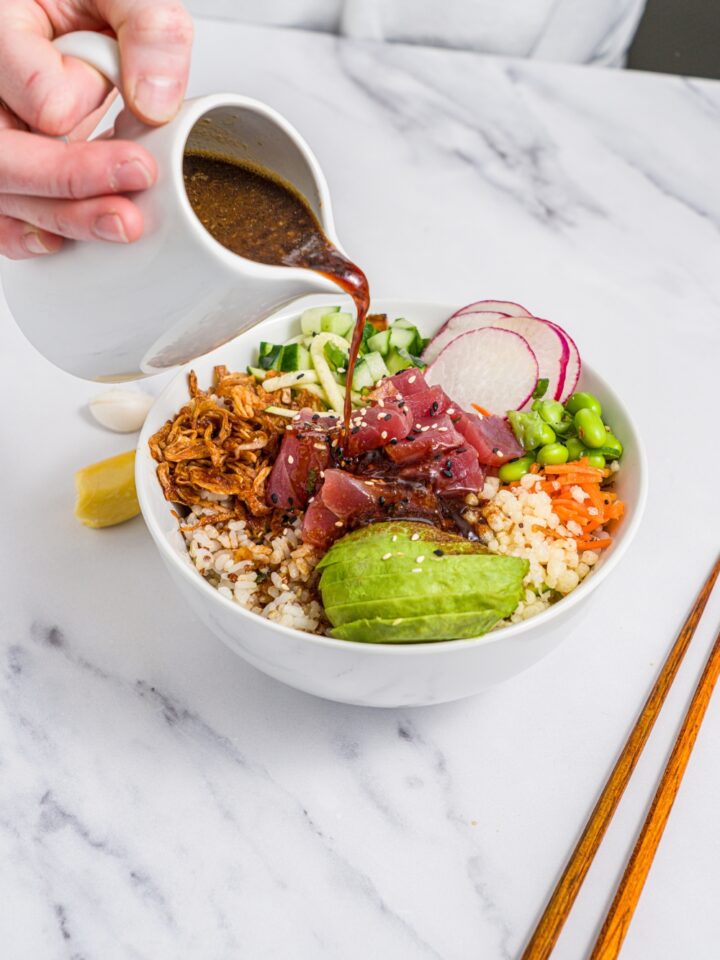 A white pitcher of poke sauce being poured over a tuna poke bowl. The bowl is served with avocado, rice, crispy onions, radishes, edamame, cucumber, and sesame seeds. The bowl is on a marble counter with a pair of chopsticks.